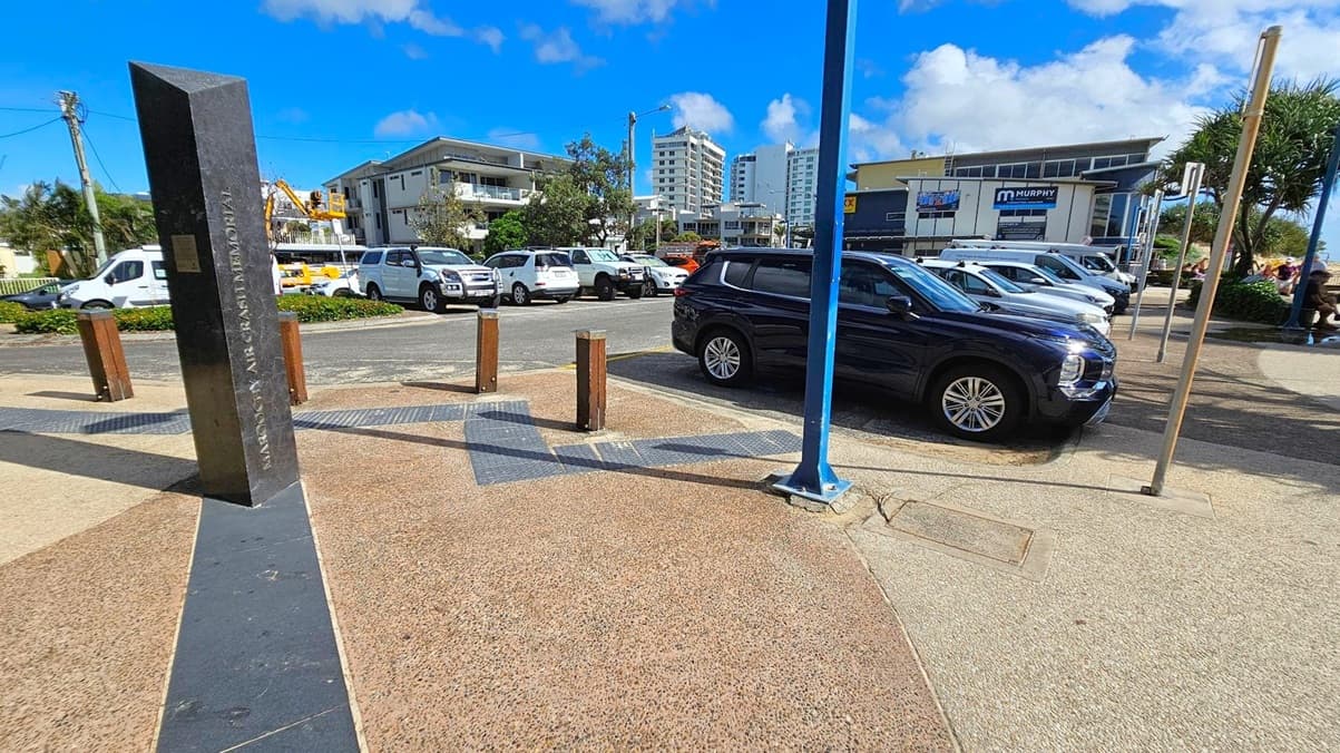 A paved area with a black obelisk on the left, inscribed with "Maroochy Air Crash Memorial". Several bollards are nearby. On the right, a row of parked cars, including a black SUV, is visible. In the background, there are buildings, including a structure with a "Murphy" sign. The sky is blue with some clouds. The ground is a mix of textured concrete and patterned tiles.
