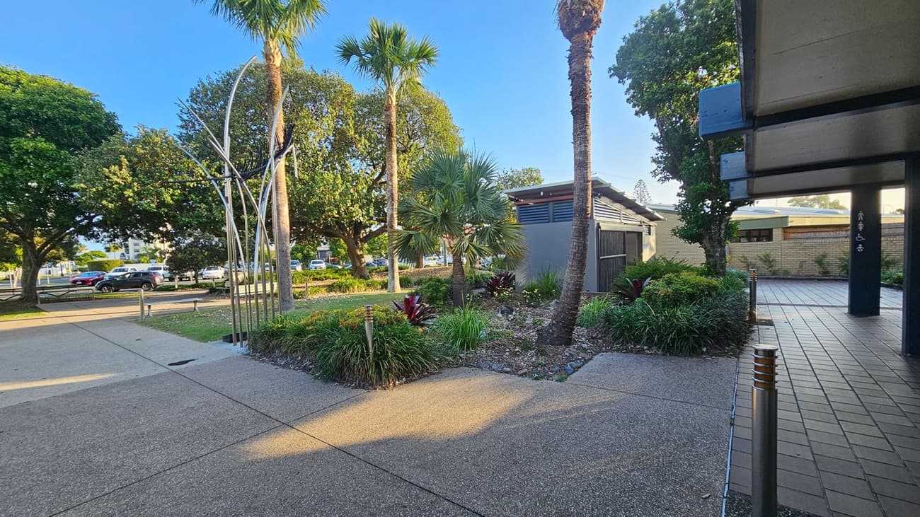 A paved walkway with a textured surface leads through a landscaped area featuring palm trees and various shrubs. On the left, a metal sculpture stands near a row of parked cars and large leafy trees. On the right, a building with a tiled entrance and a dark column is partially visible. The ground is a mix of concrete and brick paving, and the area is well-lit with small bollard lights.