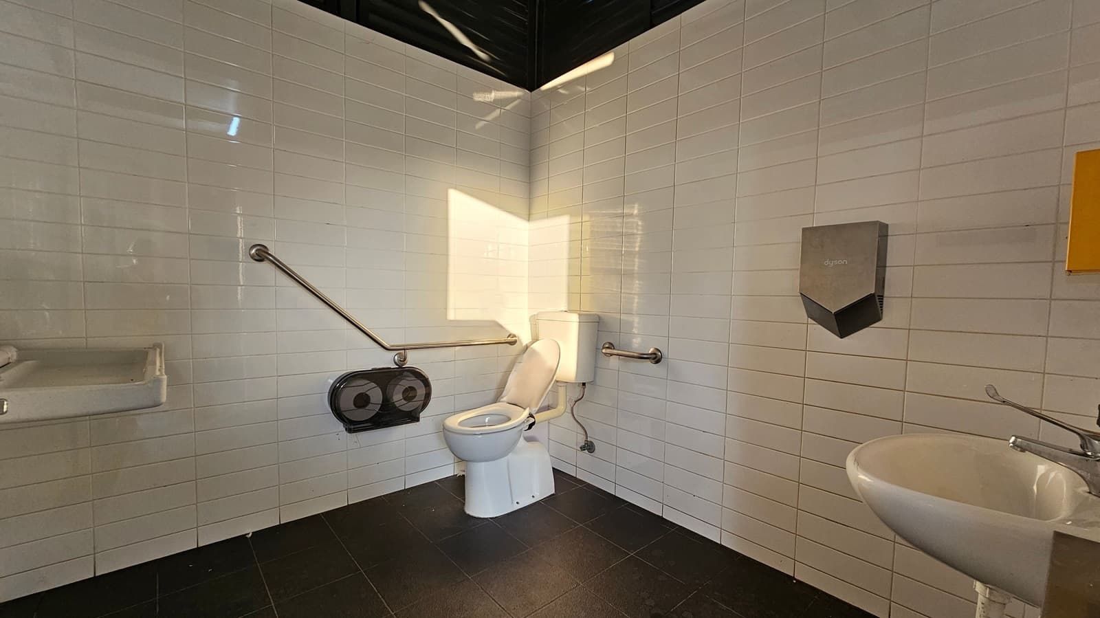 A public toilet with white tiled walls and a dark tiled floor. In the centre, there is a toilet with a white seat, equipped with metal grab bars on both sides. To the left, a sink with a soap dispenser is mounted on the wall. On the right, a hand dryer is installed above another sink. A large toilet paper dispenser is located near the toilet. The ceiling is dark, and sunlight casts a shadow on the wall.