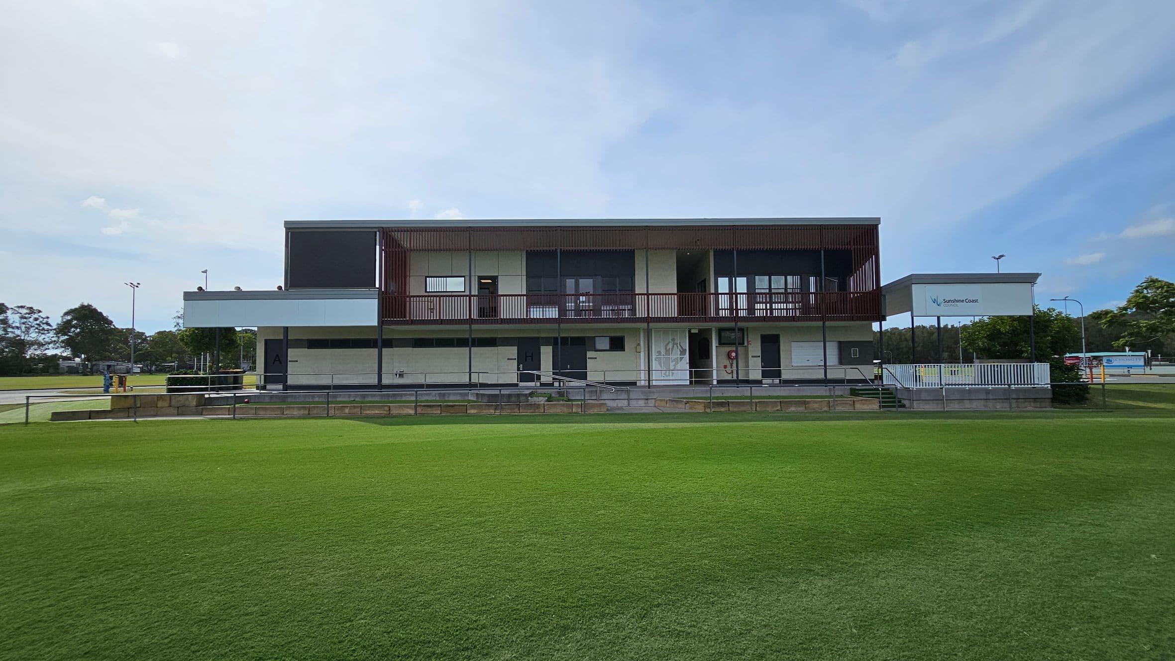 A modern, two-storey building with a flat roof and a mix of concrete and glass elements. The upper level features a balcony with railings, while the lower level has several doors and a ramp. The building is surrounded by a well-maintained grassy field. On the right, there's a sign with "Sunshine Coast" and some trees. In the background, there are more trees and a few light poles.