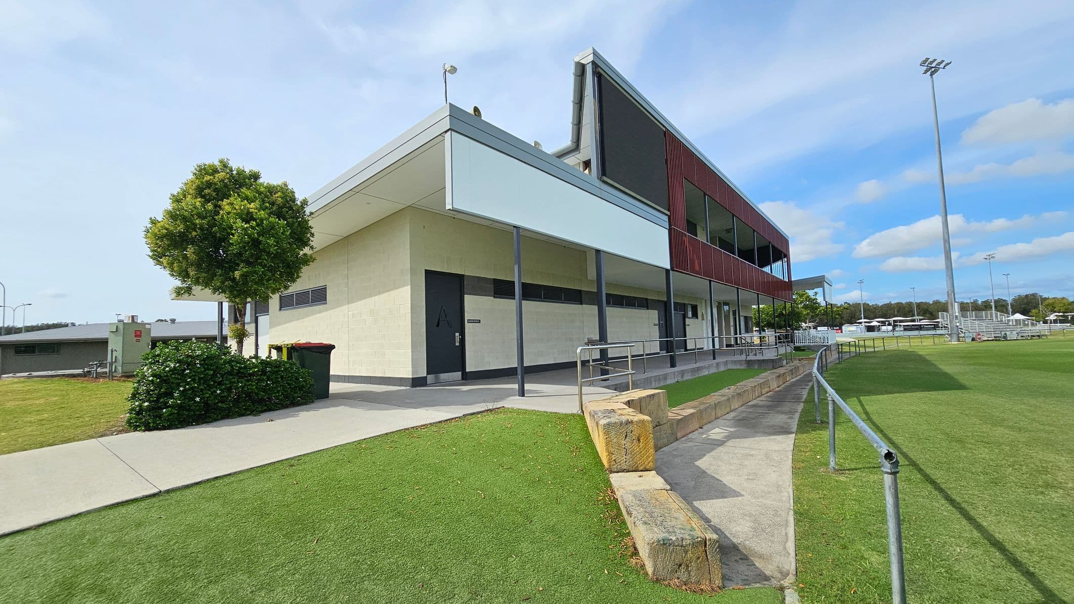 A modern sports facility with a two-storey building on the right, featuring a mix of cream and dark red panelling. A small tree and bushes are on the left near a concrete path. The foreground has a grassy area with a low stone wall and a metal railing. In the background, there are sports fields with tall floodlights and several white structures. The sky is clear with some clouds.