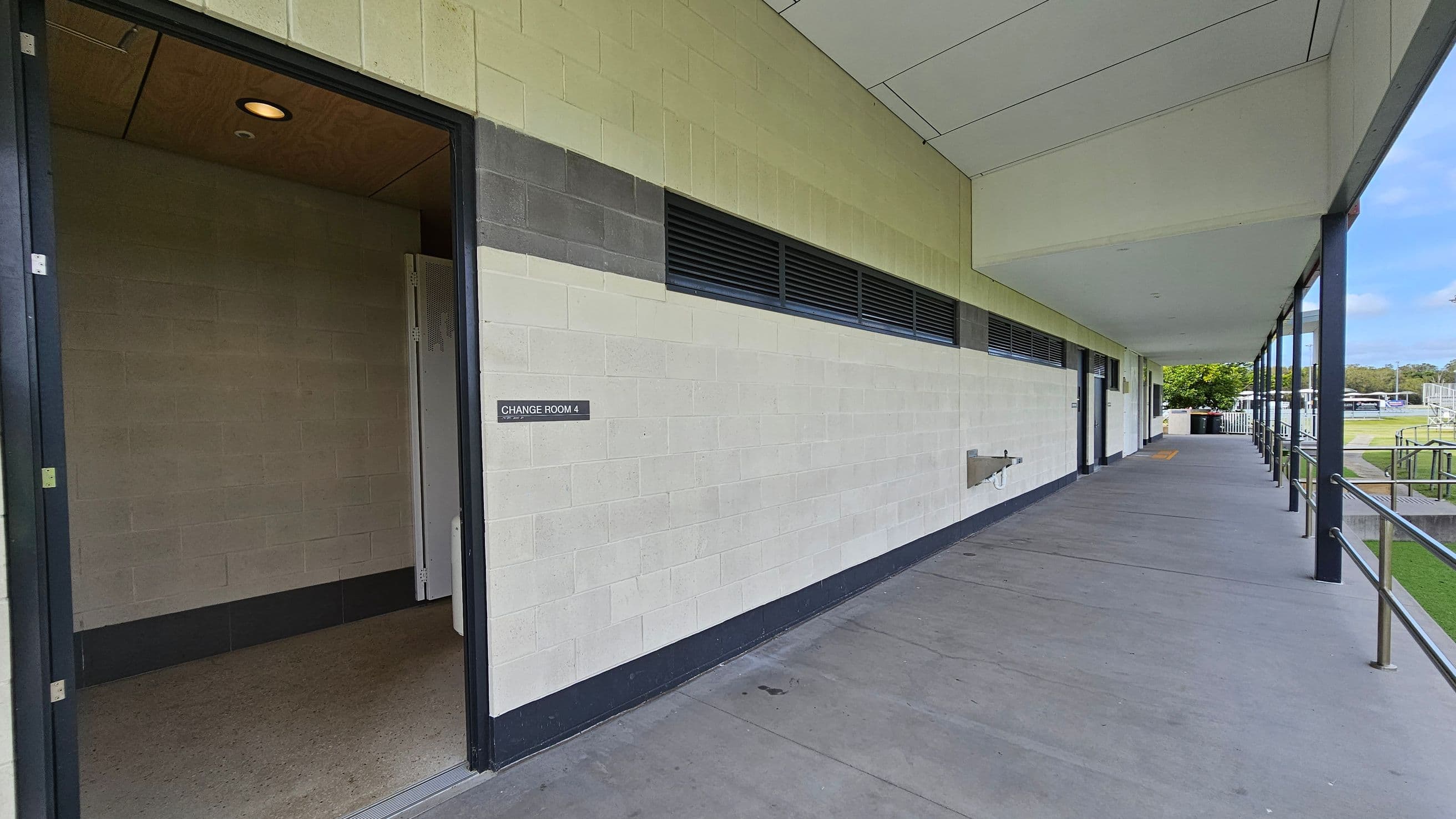 A corridor with a concrete floor and a ceiling with panels. On the left, there's an open door labelled "Change Room 4" leading into a room with a light-coloured floor. The wall is painted in light green and grey with ventilation panels. On the right, a railing runs along the edge of the corridor, and beyond it, there is a grassy area with trees and outdoor structures. The corridor extends into the distance with more doors and a water fountain visible.