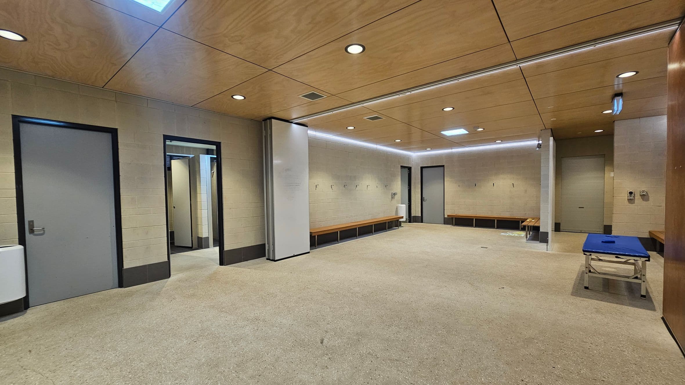 A spacious locker room with beige walls and a light brown ceiling featuring recessed lighting. On the left, there are two grey doors and a white wall-mounted unit. In the centre, a doorway leads to another area. Along the right wall, wooden benches are fixed, and a blue padded table is positioned nearby. The floor is covered with a light-coloured carpet.