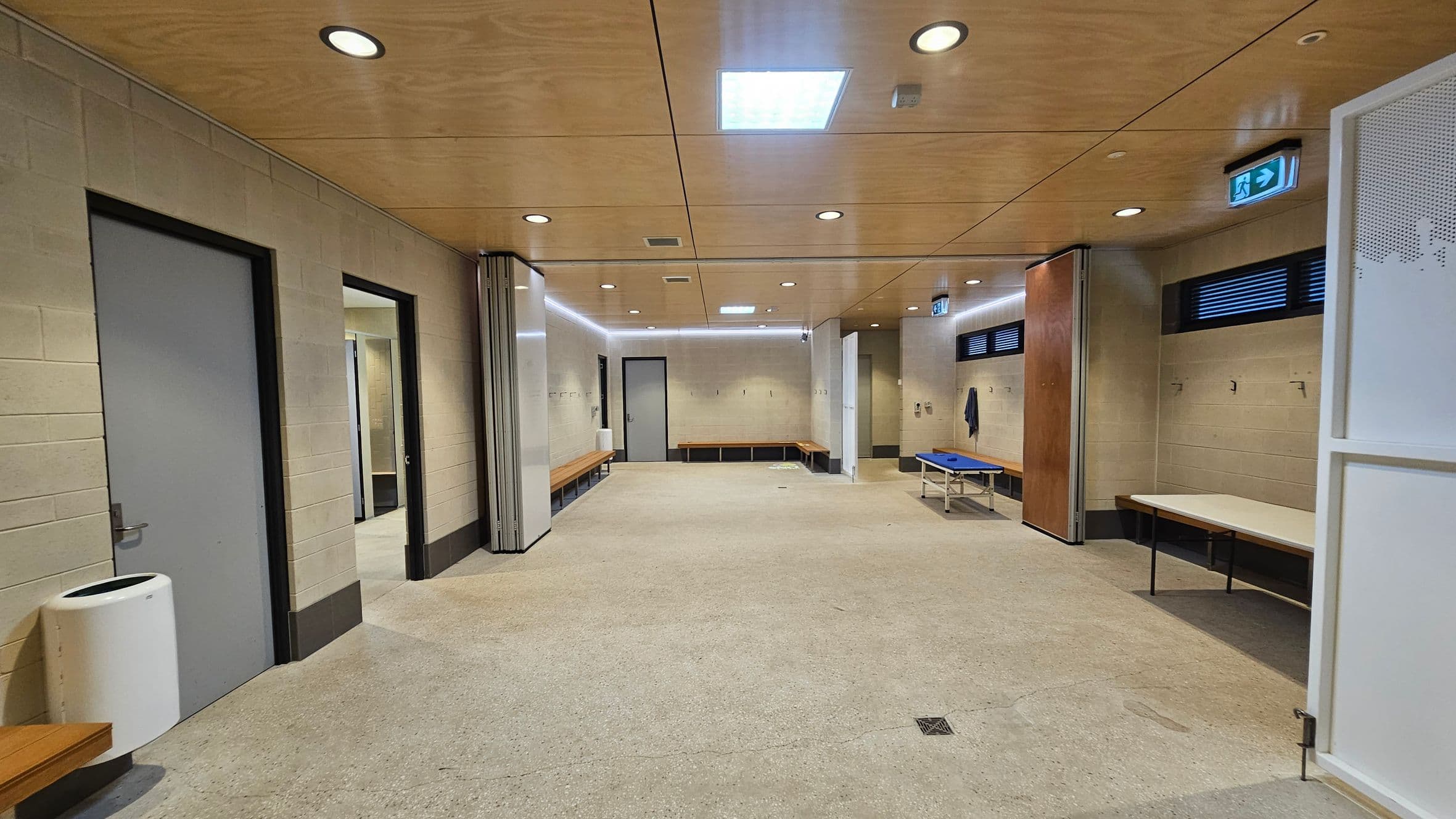 A spacious locker room with beige walls and a light-coloured ceiling featuring recessed lighting. On the left, there are grey doors and a white bin. Benches line the walls, with hooks above them. On the right, a blue bench and a table are visible. The floor is a speckled light grey. An emergency exit sign is above a door on the right.