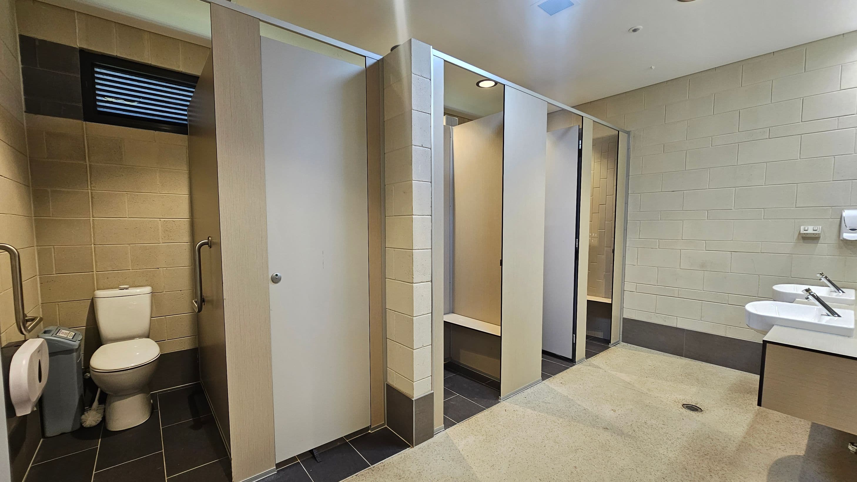 A public restroom with beige tiled walls and a light-coloured ceiling. On the left, there is a toilet cubicle with a white toilet, grab bars, a toilet paper dispenser, and a sanitary bin. The middle section has several closed cubicle doors. On the right, there are two white sinks with silver taps mounted on a countertop, and a soap dispenser on the wall. The floor is a mix of dark tiles and light-coloured flooring.