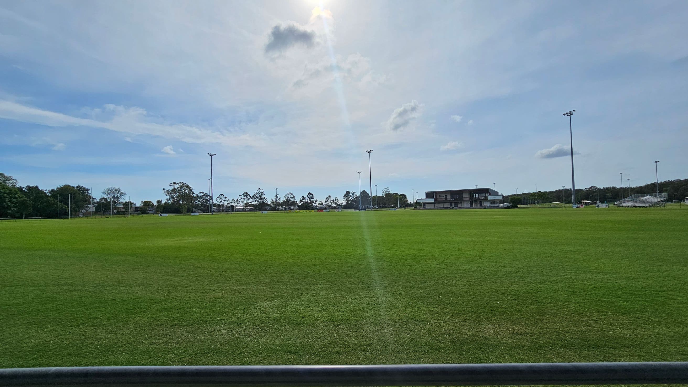 A large, open grassy field with a few patches of brown. In the background, there are several tall floodlights and a building to the right. Trees line the horizon, and there are some small structures or equipment scattered across the field. The sky is mostly clear with a few clouds.