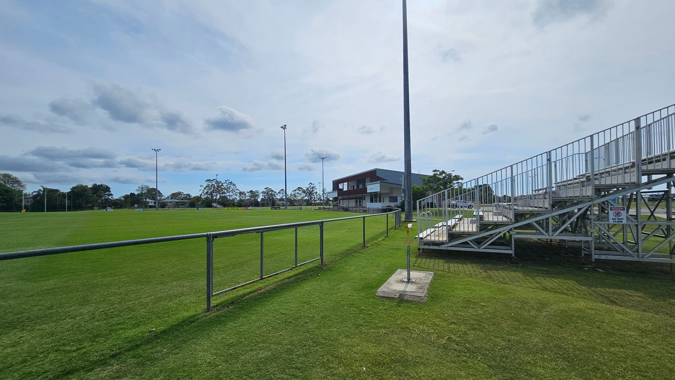 A sports field with a well-maintained grass surface. On the right, there are metal bleachers with a sign that reads "DO NOT CLIMB." In the centre, a tall floodlight pole stands near a building with a balcony. The background features trees and additional floodlights. A metal railing runs along the left side of the field.