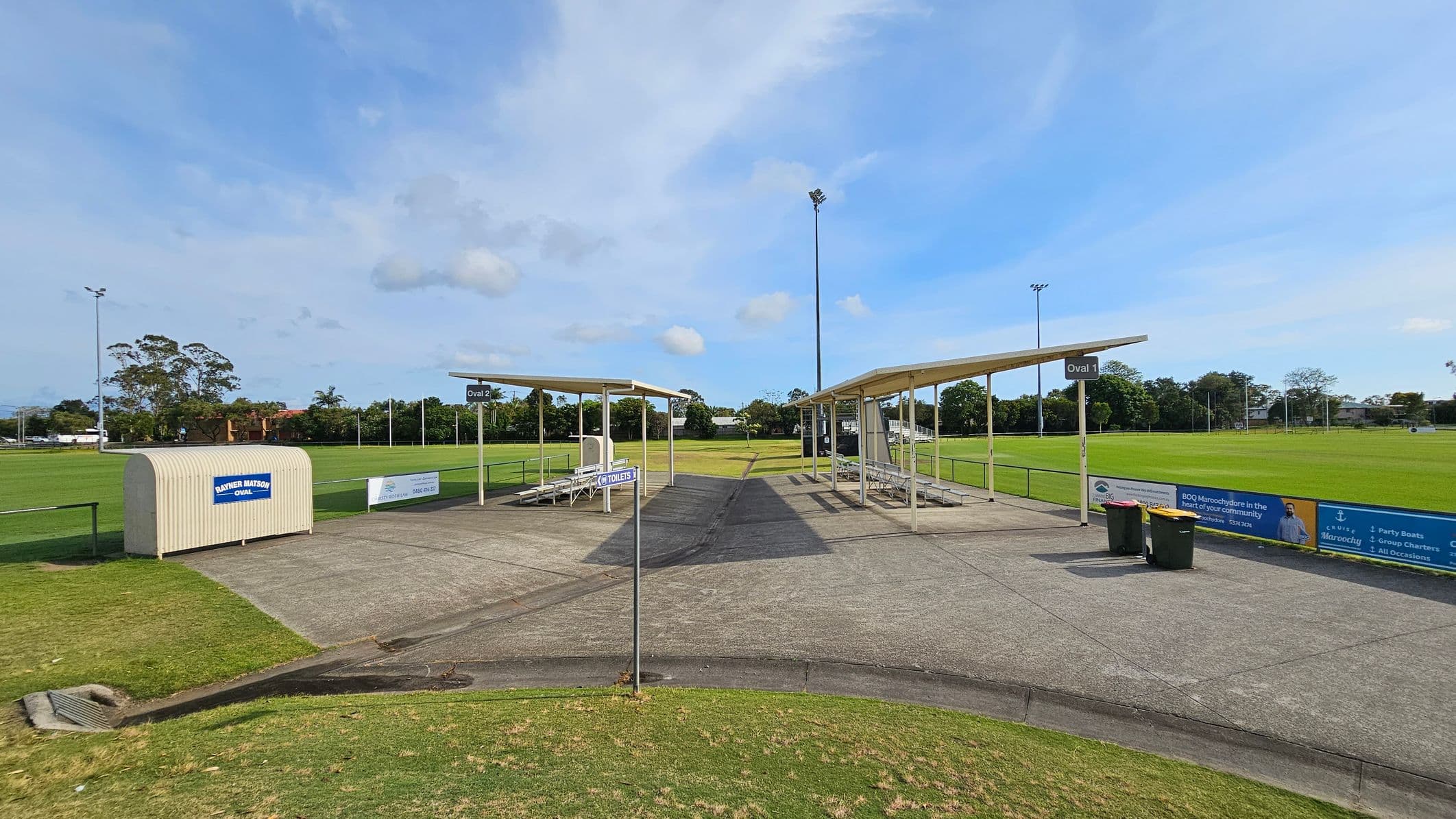 A sports field with two covered seating areas in the centre, labelled "Oval 1" and "Oval 2". The ground is a mix of concrete and grass. A small structure on the left has a sign reading "Panther Match Day Quarters". A signpost indicates the direction to toilets. Two bins are near the seating on the right. The background features a grassy field with goalposts and trees. Advertising banners line the fence on the right.