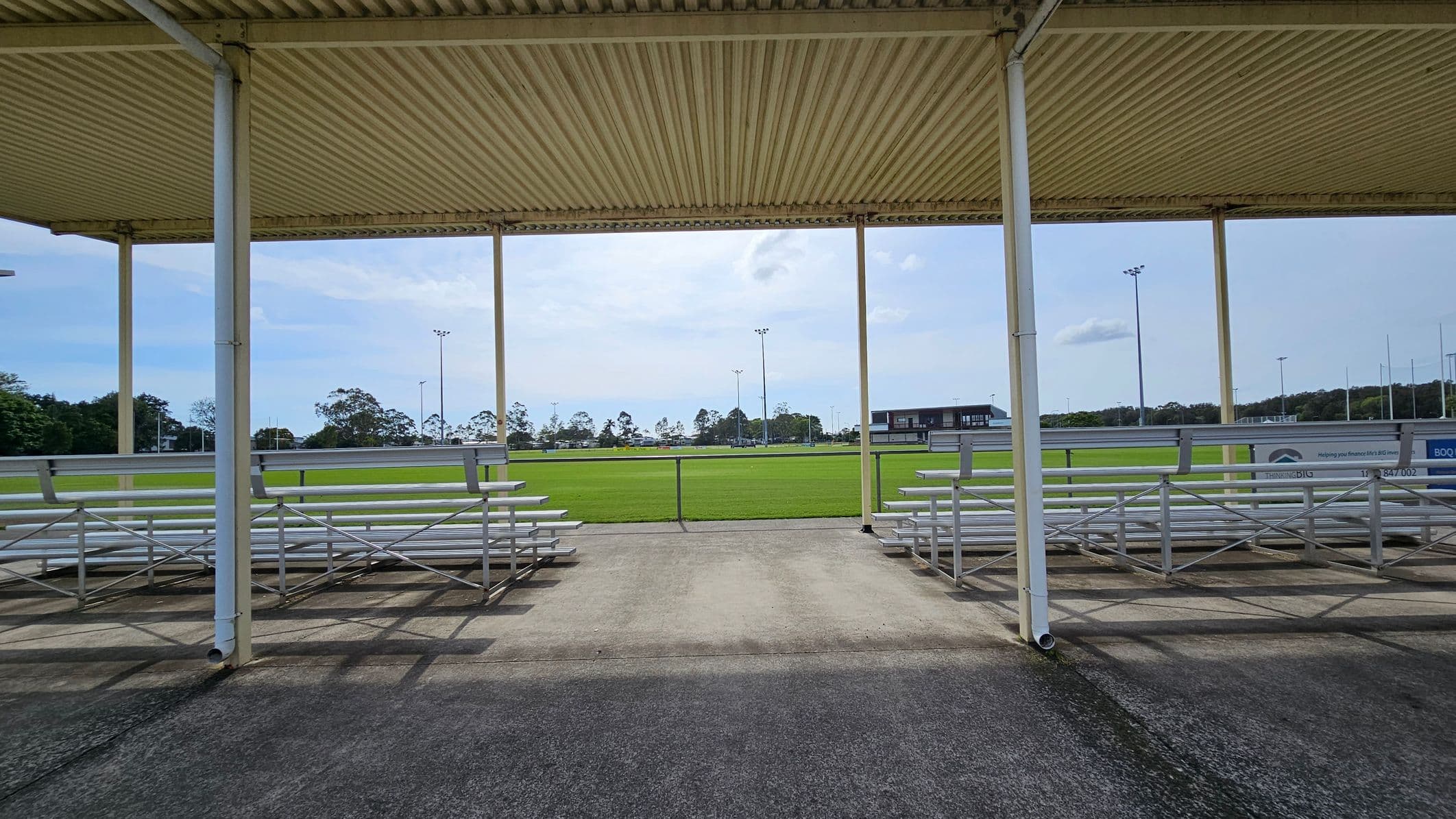 A sports field with a green grass pitch in the background, surrounded by tall floodlights. In the foreground, there are metal bleachers on both the left and right sides, under a corrugated metal roof supported by white poles. The ground is paved with concrete, and there are trees and a building visible in the distance.