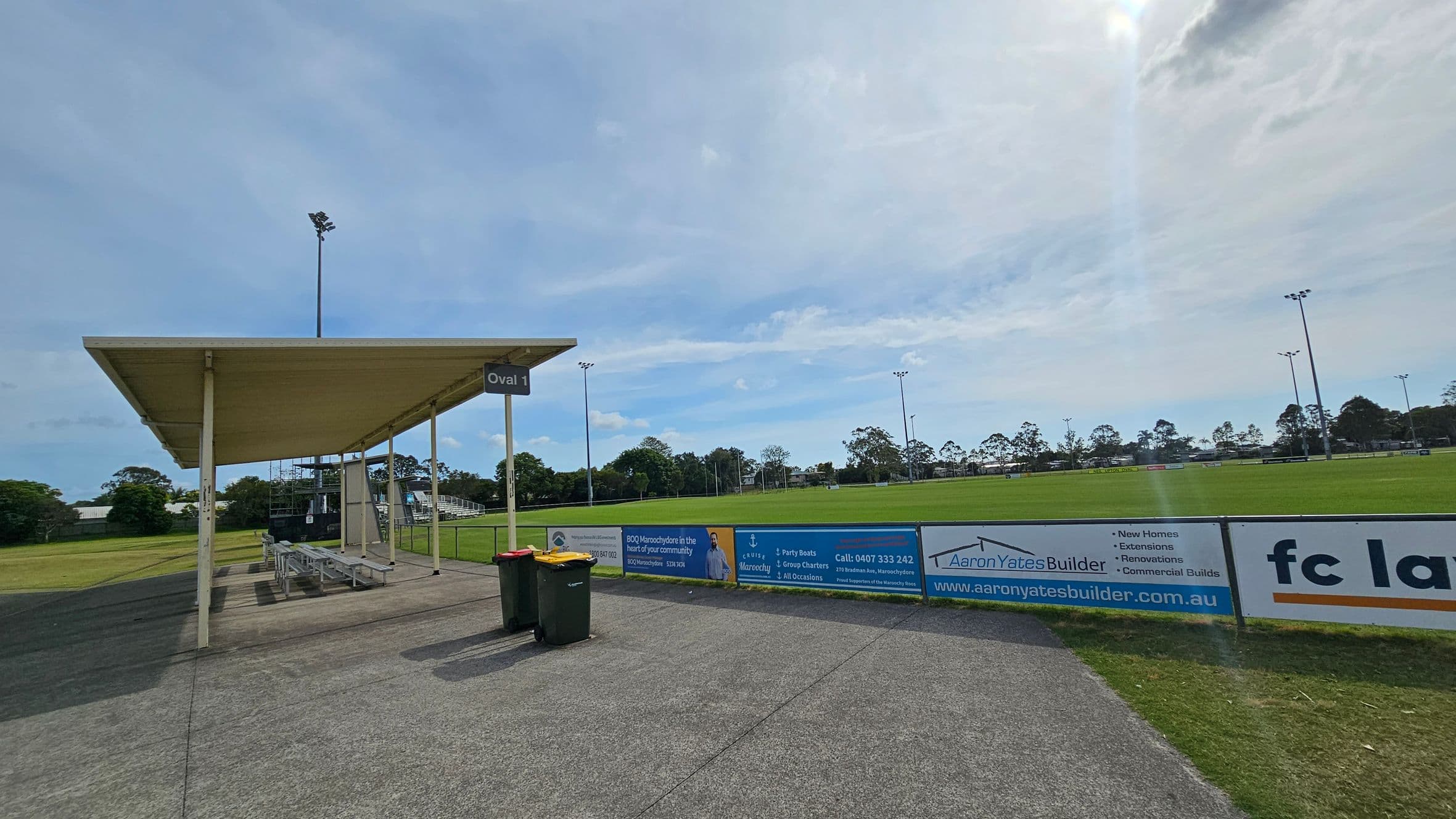 A sports field with a green grass pitch is in the centre, surrounded by a low fence with advertising banners. On the left, there's a covered seating area with benches and a sign reading "Oval 1". Two rubbish bins are placed nearby on a paved path. Tall floodlights are visible around the field, and trees line the background. The sky is overcast.