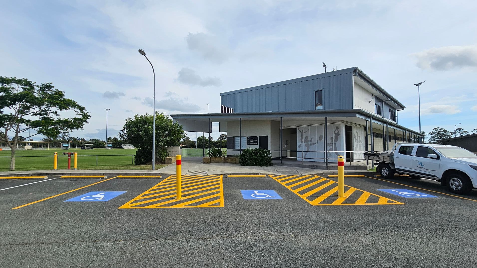 A modern building with a grey exterior is on the right, featuring large windows and a covered entrance. In front, there are marked disabled parking spaces with blue symbols and yellow lines. A white utility vehicle is parked in one of the spaces. On the left, there is a grassy field with a few trees and a tall streetlamp. The background includes more greenery and sports fields. The ground is paved, and there are yellow bollards near the parking area.