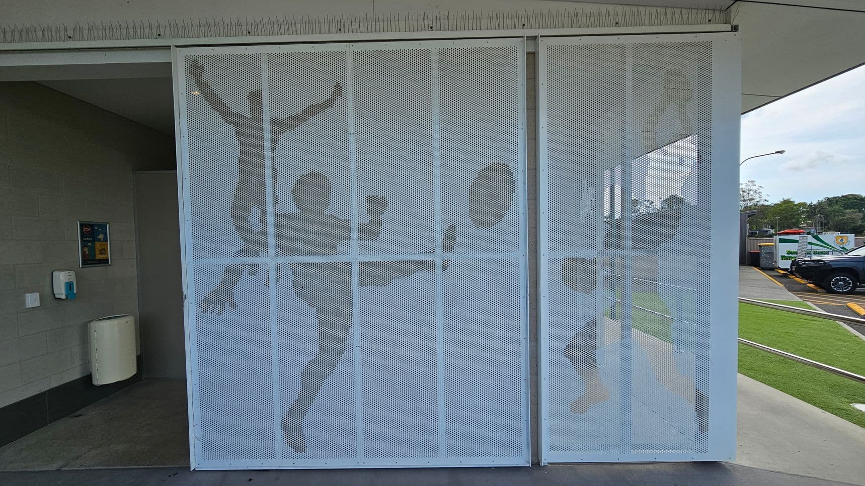 A perforated metal screen with silhouettes of people engaged in athletic activities, such as kicking a ball, is positioned in the centre. On the left, there's a wall with a hand sanitiser dispenser and a waste bin. The right side shows a parking area with a black vehicle and some greenery. The ceiling has a row of anti-bird spikes.