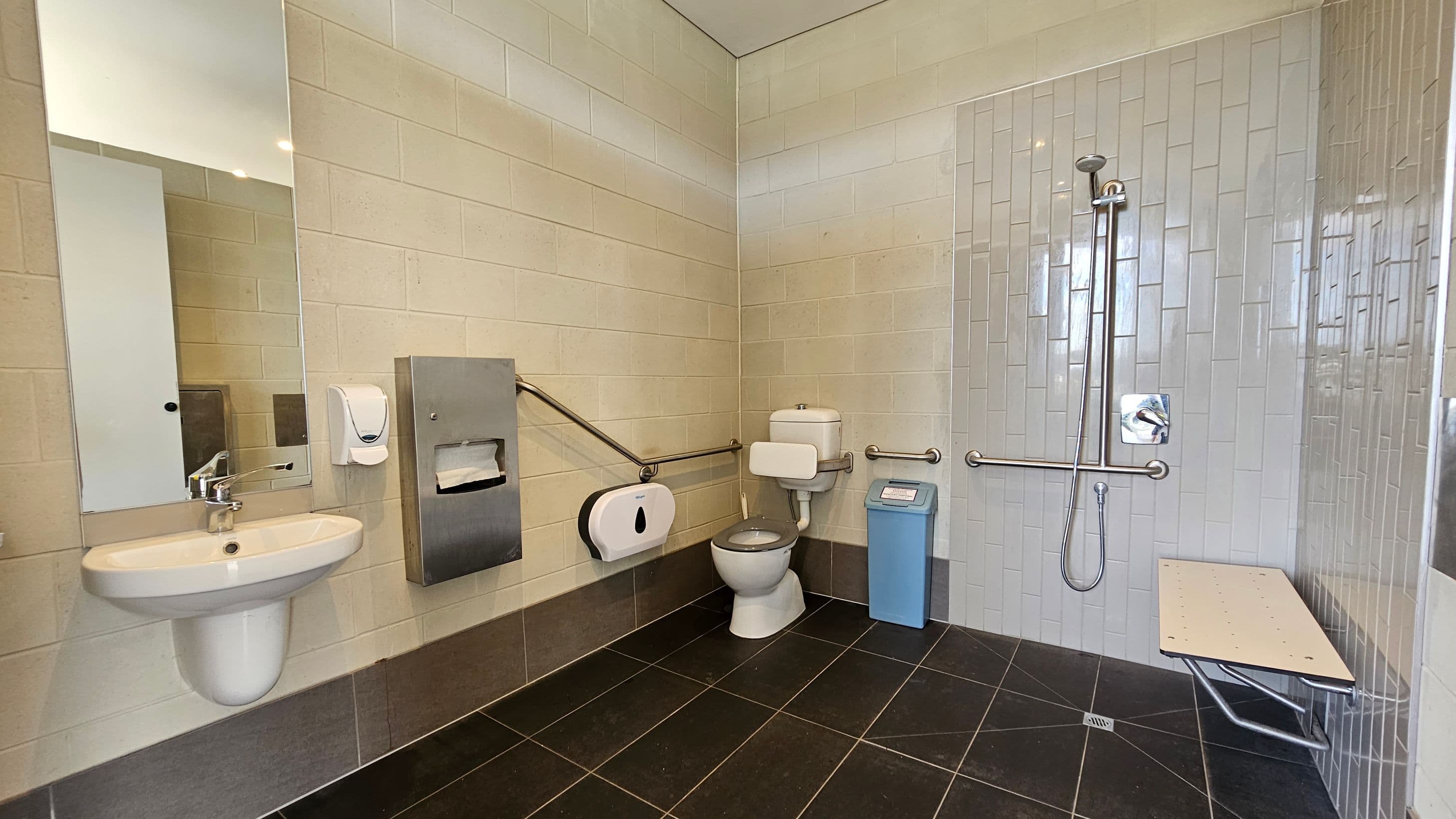 Accessible bathroom with a white sink and mirror on the left. A soap dispenser and paper towel holder are mounted on the wall. In the centre, a toilet with support rails and a blue bin nearby. On the right, a shower area with a handheld showerhead and grab bars, alongside a foldable bench. The walls are tiled in light colours, and the floor has dark tiles.