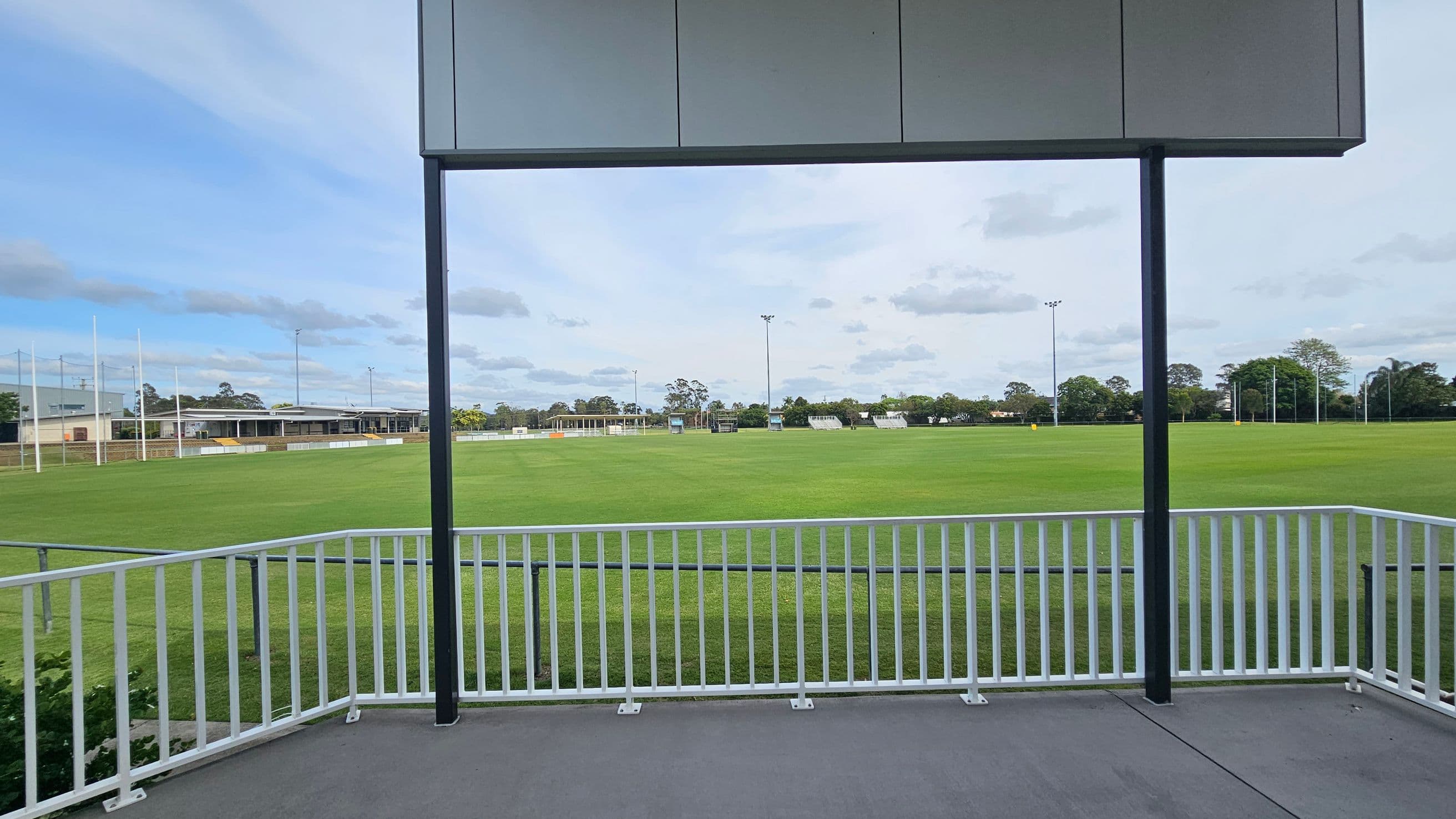 A sports field with a well-maintained green lawn is visible, surrounded by a white railing in the foreground. In the background, there are goalposts and a building with a flat roof on the left. Trees and additional goalposts are scattered along the horizon. A metal structure with a roof is partially visible at the top of the image.