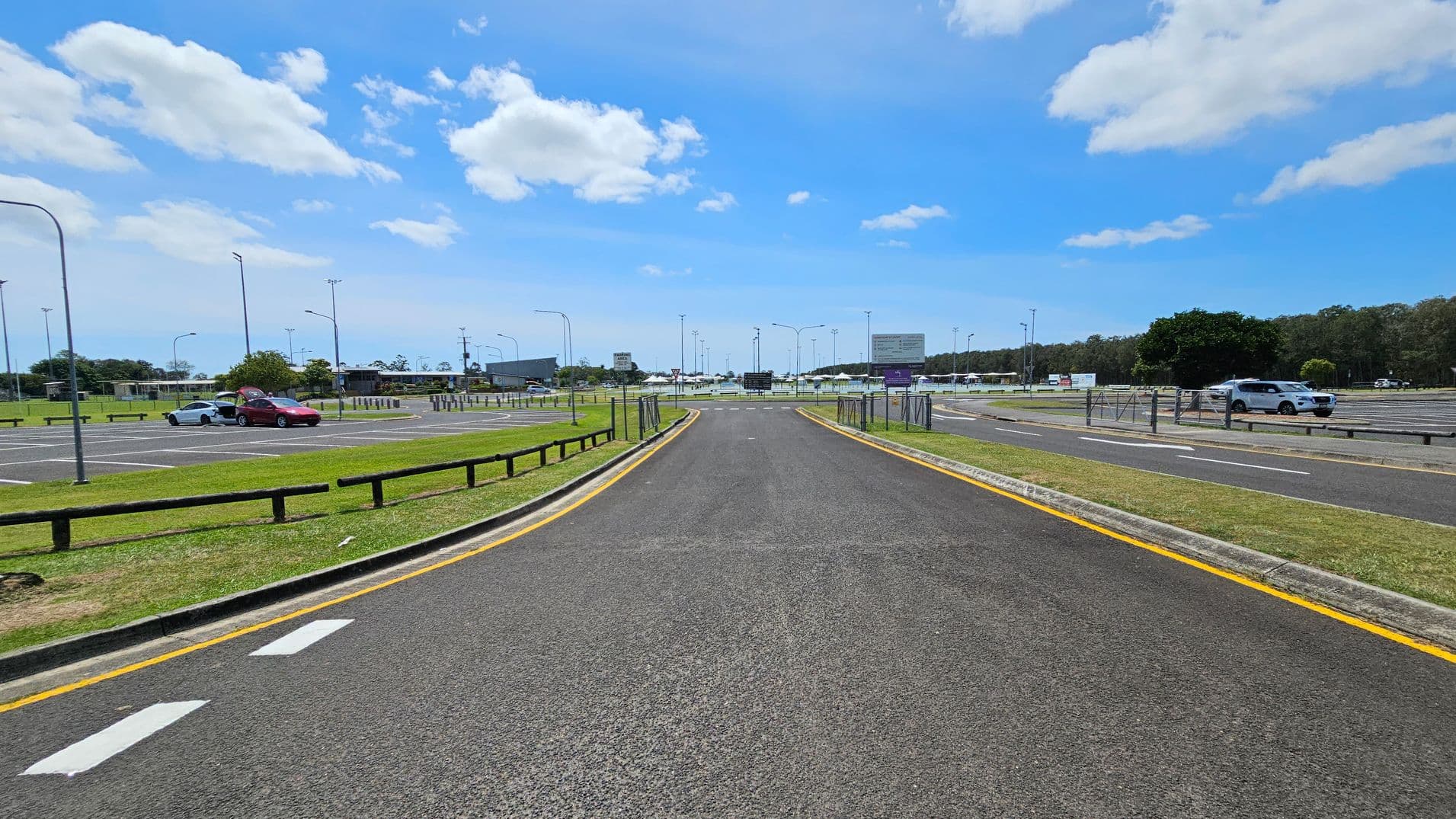 A road curves to the left with a yellow line marking its edge. On the right, a blue sign reads "Maroochydore Multi Sports Complex." Beyond the road, there are parking spaces with a few cars and a grassy area with trees in the background. Several streetlights and signs are visible along the road. The sky is clear with a few clouds.