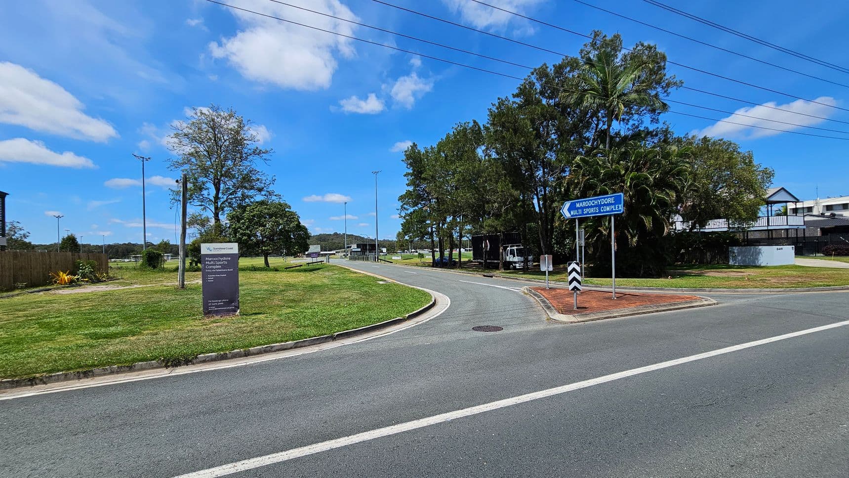 A road curves to the right, bordered by green grass and trees. On the left, a sign reads "Maroochydore Multi Sports Complex" with additional details. A blue directional sign points towards the complex. Trees and a few buildings are visible in the background. Overhead, power lines stretch across the scene. The road surface is grey with white markings.