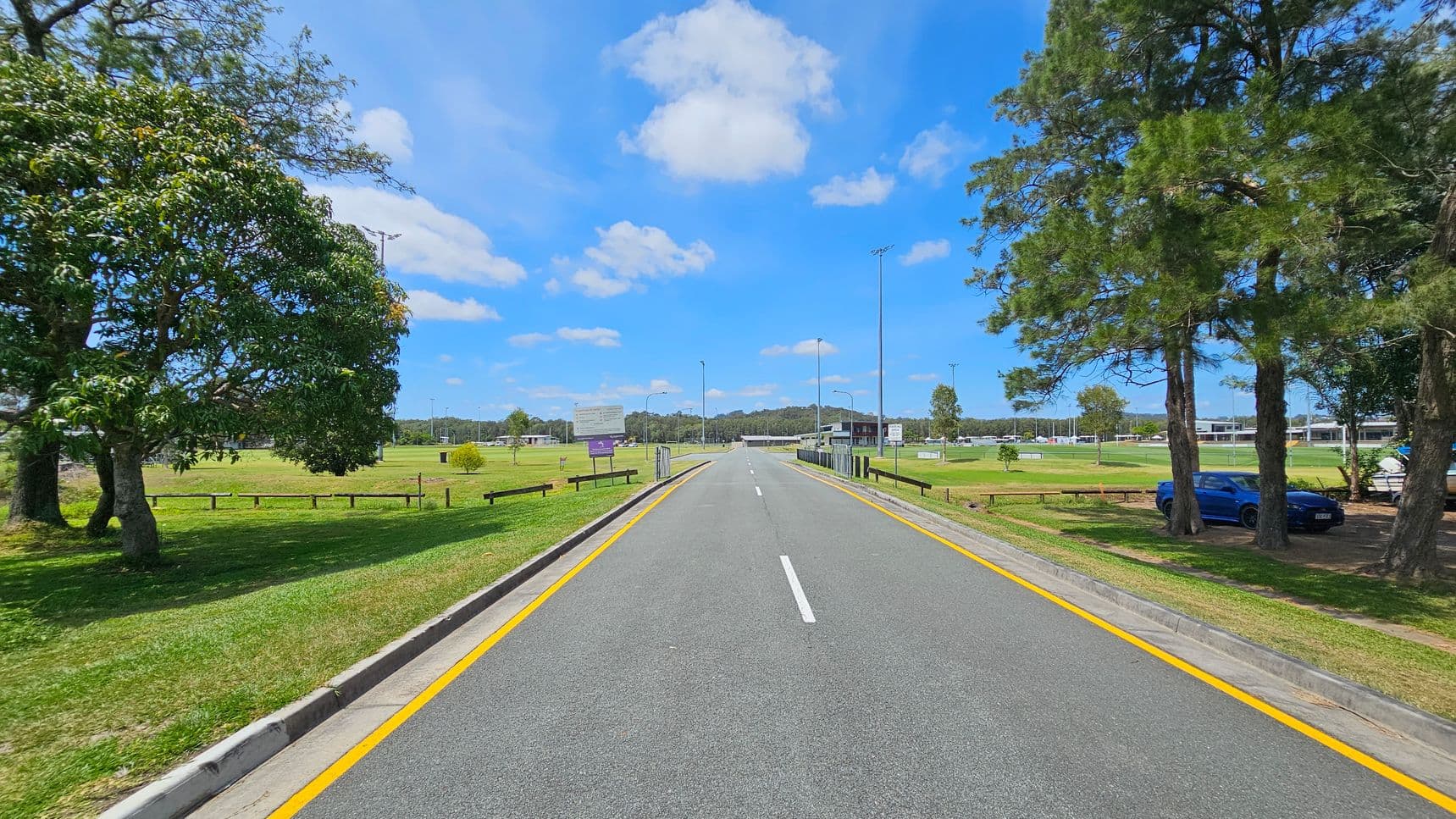 A straight road with yellow lines runs through the centre, flanked by grassy areas. On the left, there are several large trees and a wooden fence. On the right, more trees are visible, with a blue car parked under them. In the background, there are sports fields with goalposts and a few buildings. The sky is clear with scattered clouds.