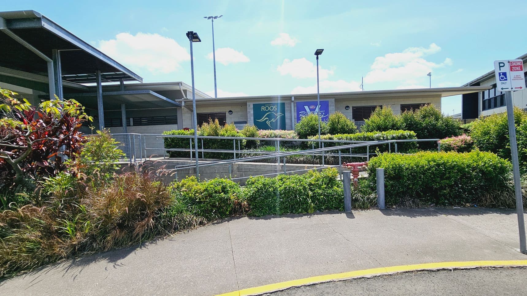 A building with a flat roof is in the centre, featuring a covered entrance with steps leading up to it. In front, there are several marked disabled parking spaces on an asphalt surface. To the left, a grassy area with a tree and two bins is visible. On the right, there are bushes and a signpost. The background shows a sports field with goalposts and more greenery.
