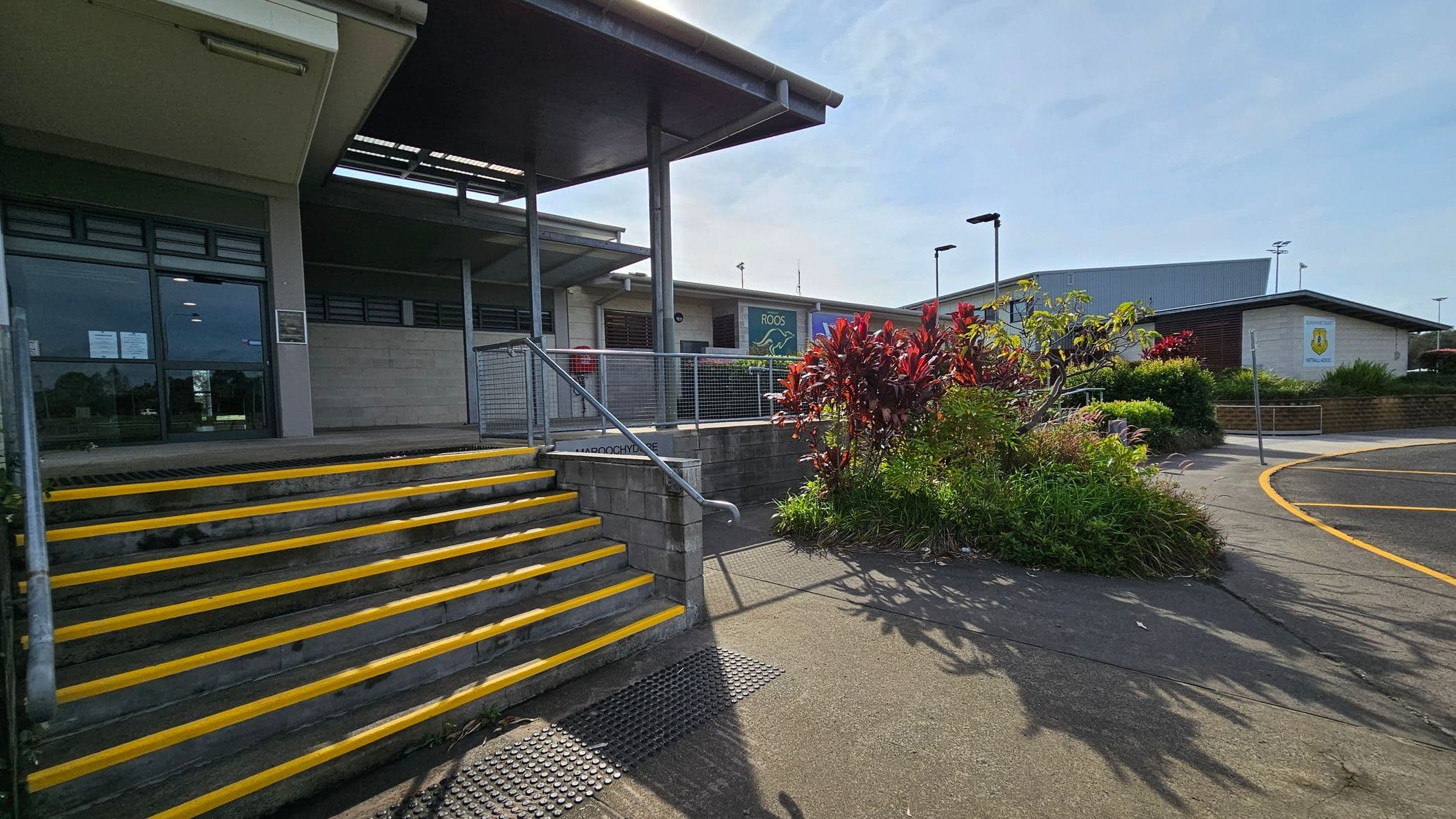 Concrete steps with yellow edges lead up to a building entrance on the left, featuring large glass windows and a metal railing. In the centre, a garden area with red and green plants is bordered by a concrete path. On the right, a curved road with yellow markings is visible. The building has a flat roof and a sign with a logo on the wall. The area is well-lit, and there are street lamps in the background.