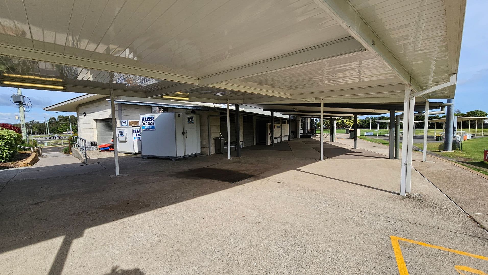 A covered walkway with a white ceiling and supporting poles. On the left, there's a building with a ramp leading to a door, and signs for "Kleer Cold Room Hire" and "Kleer Ice." A large white cold room unit is visible. The ground is concrete, and there are shadows from the structure. In the background, there is a grassy area with trees and a road.