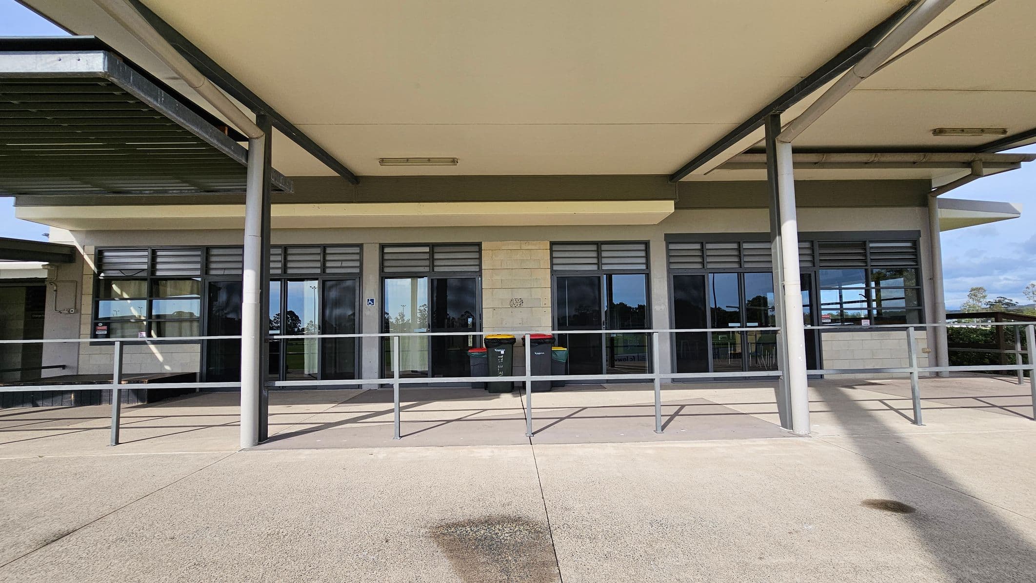 A building with a flat roof supported by metal columns. The facade features large glass windows with louvres above them. In front of the windows, there is a metal railing and several bins with coloured lids. The ground is paved with concrete, and there is a shadow cast by the roof. The background shows some greenery and a partly cloudy sky.