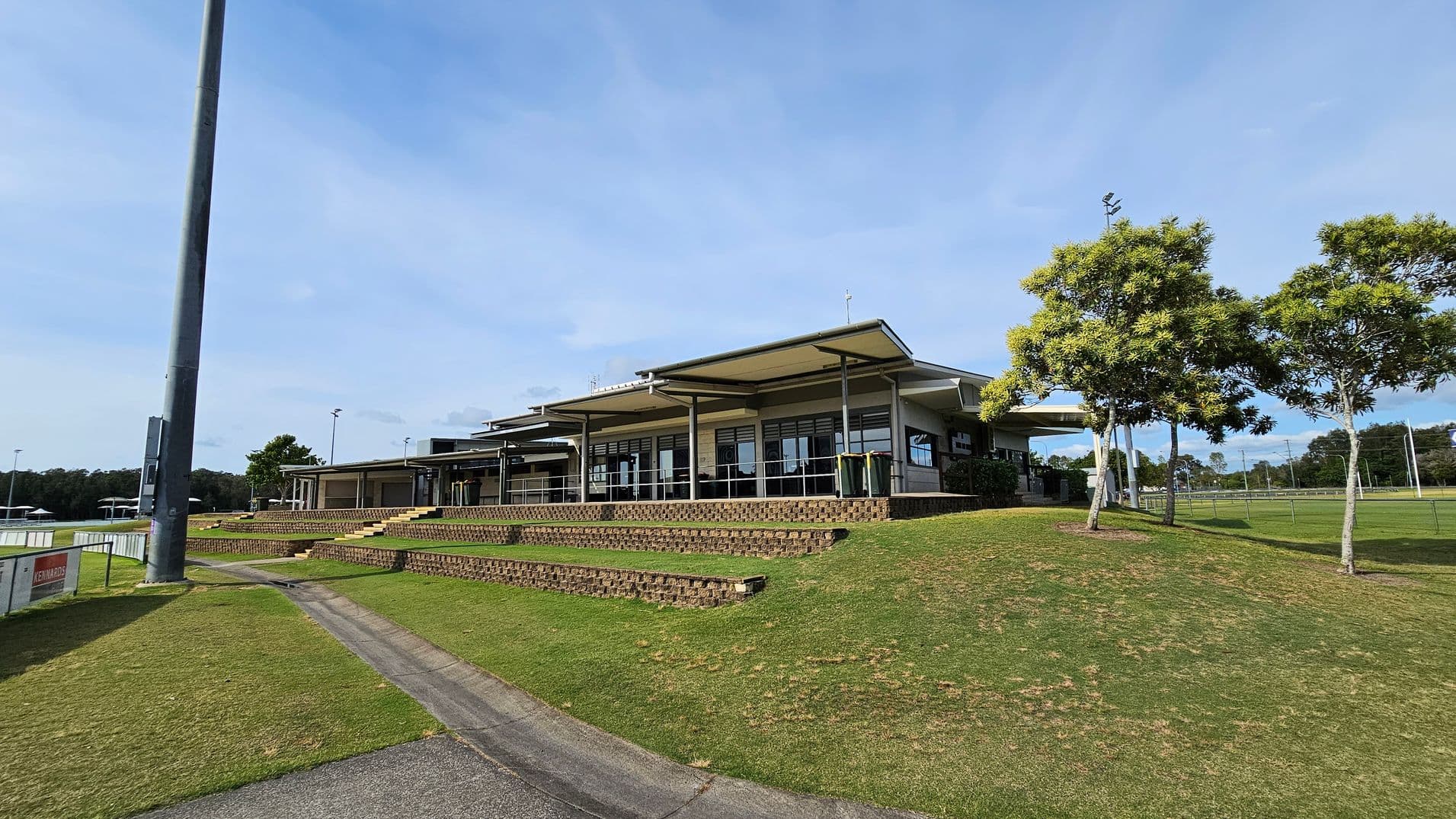 A modern building with a flat roof and large windows is situated on a grassy hill. In the foreground, there are tiered brick steps leading up to the building. To the right, several trees with green foliage are visible. On the left, a tall light pole stands near a pathway that runs alongside the grass. In the background, there are sports fields with goalposts and more trees.