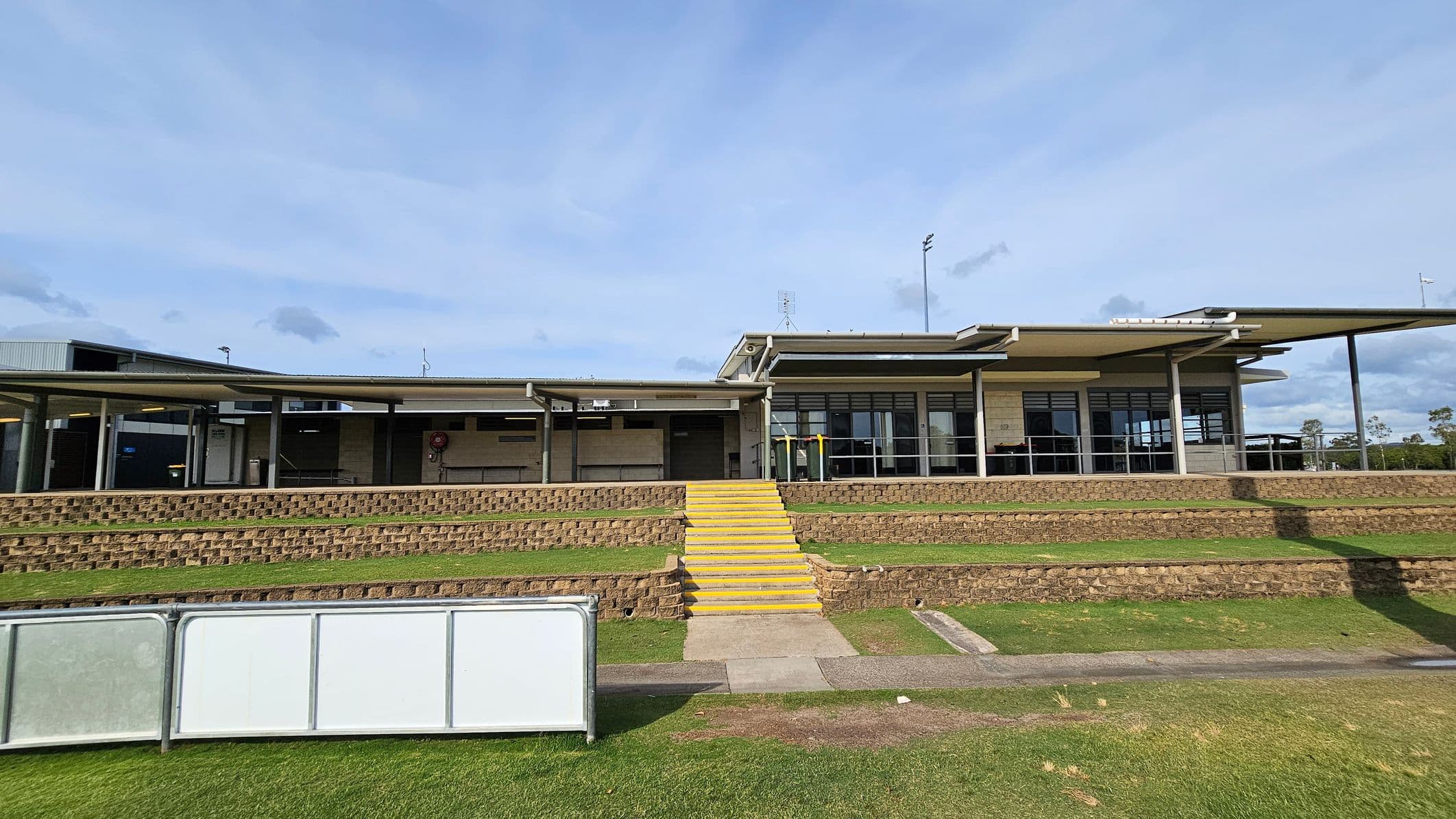 A modern building with a flat roof and large windows is in the centre. It has a series of yellow steps leading up to it, flanked by tiered brick retaining walls. The foreground features a grassy area with a concrete path and a white barrier. The building has a covered walkway supported by columns, and there are some trees visible in the background.