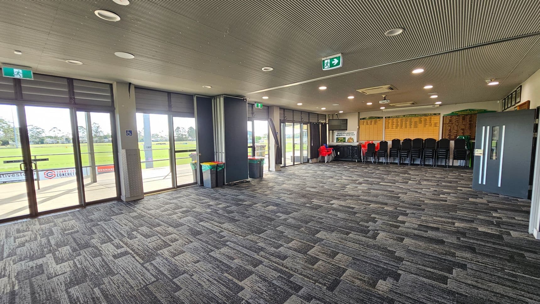 A spacious room with large glass doors on the left, offering a view of a grassy field outside. The floor is covered with patterned grey carpet tiles. On the right, there are stacked black chairs and a few red chairs near a table. The walls display wooden plaques. The ceiling is lined with recessed lights and features a grey, textured finish. An exit sign is visible above the doors.