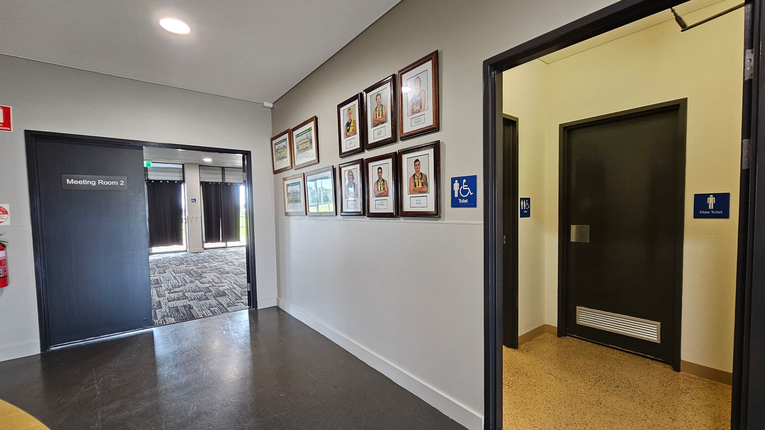 A hallway with a dark floor and light walls. On the left, there's a door labelled "Meeting Room 2" with a fire extinguisher nearby. The middle wall displays several framed photographs. On the right, there are two doors, one marked with a sign for an accessible toilet and the other for a male toilet. The ceiling is plain with a circular light fixture.