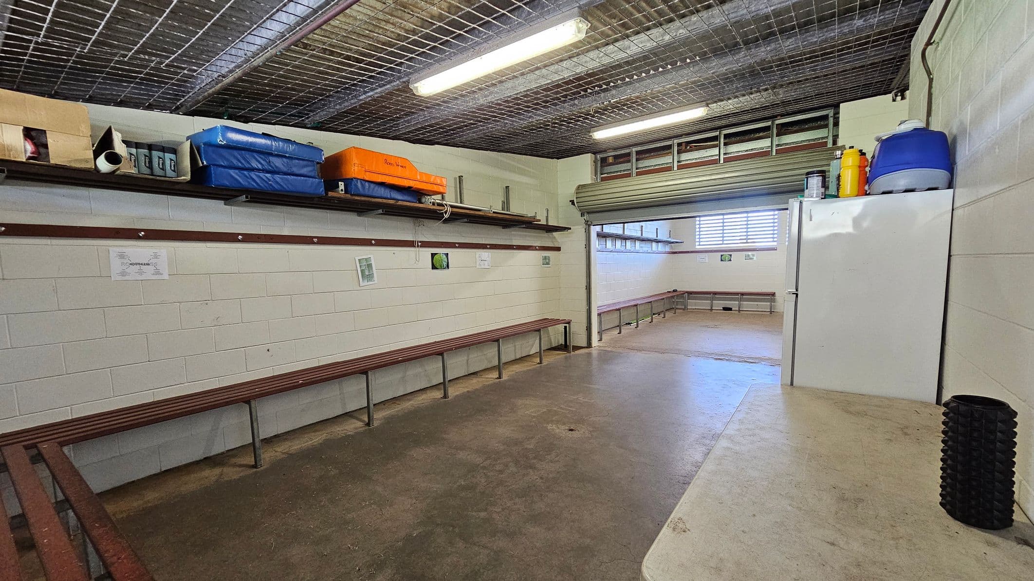 A room with concrete floors and a wire mesh ceiling. On the left, there are wooden benches along the walls and shelves above holding various items, including blue and orange mats. On the right, a white refrigerator with a blue cooler and yellow container on top. A black cylindrical object is on a nearby surface. The back wall has a partially open roller door and a window with horizontal bars, allowing light into the space.