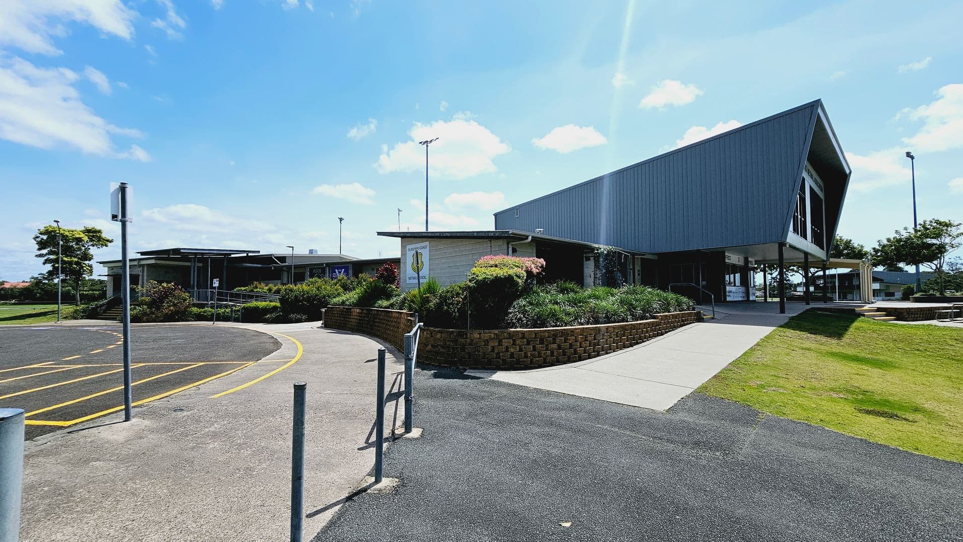 A modern building with a sign reading "Sunshine Coast Netball Association" is centred in the image. It has large windows and a flat roof. In front, there are several rows of metal picnic tables on tiered brick platforms. The foreground features a well-maintained grassy area with a concrete path leading to the building. On the left, there are power poles and a car park, while a large tree is visible on the right.