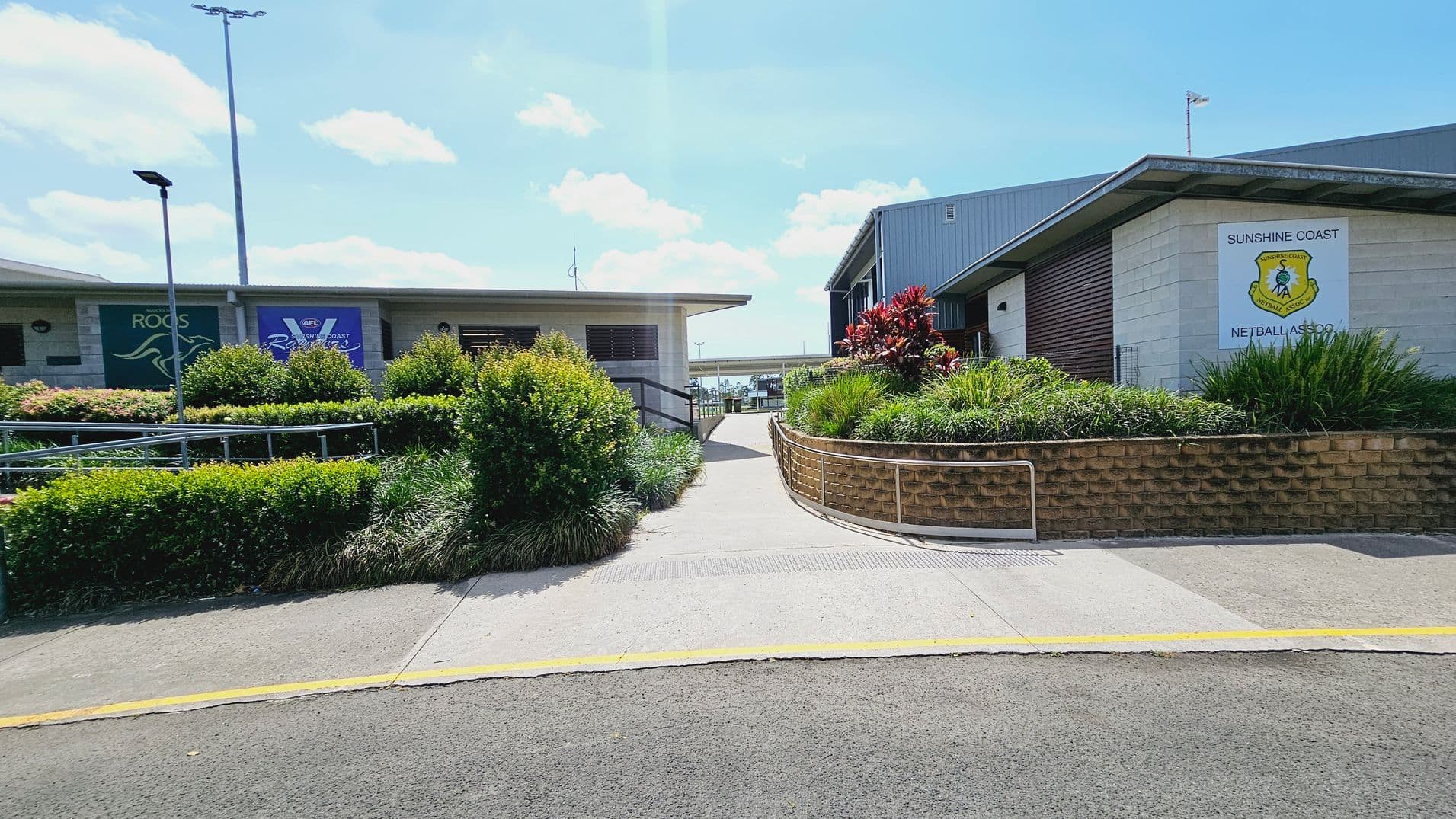 A pathway leads between two buildings. On the left, a building displays signs for "ROOS" and "AFL Sunshine Coast." On the right, another building has a sign for "Sunshine Coast Netball Association." Both buildings are surrounded by well-maintained greenery, including bushes and red-leaved plants. The path is bordered by a low brick wall and metal railing. The ground is paved, and there are tall light poles in the background.