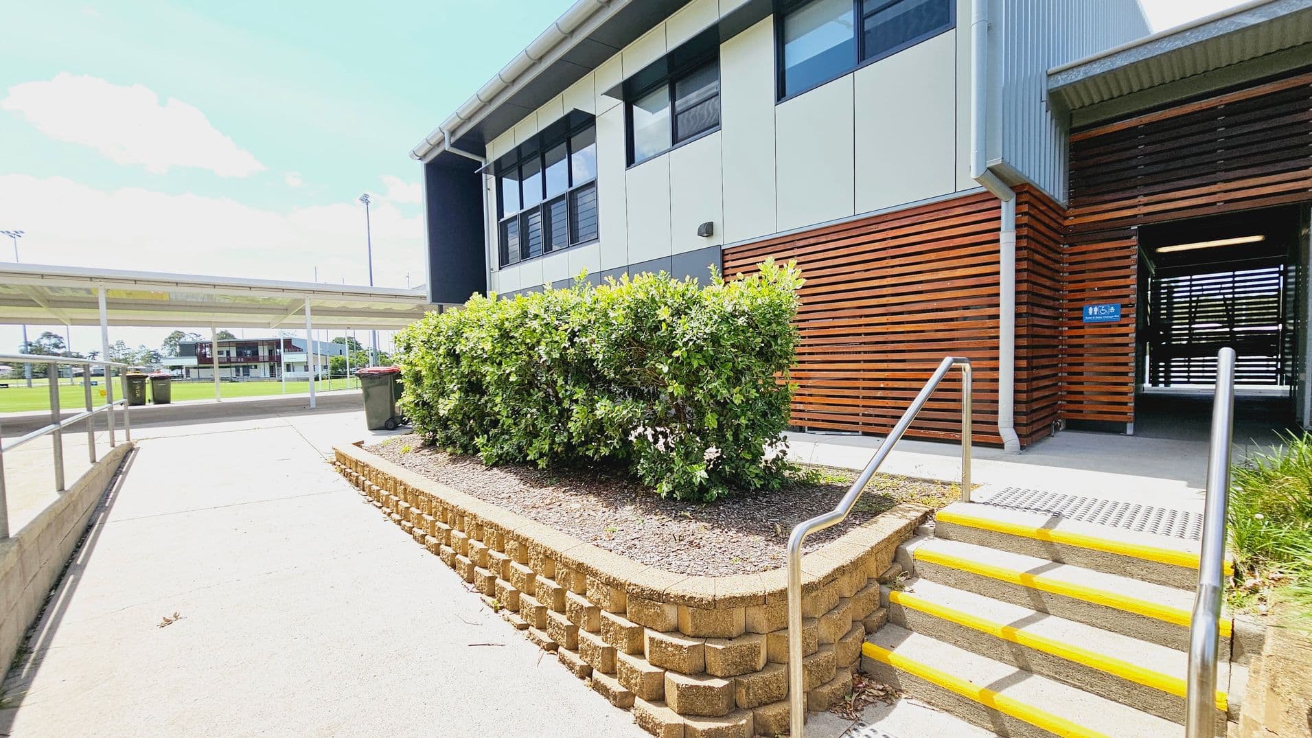 A modern building with a mix of metal and wooden panelling on the right. A set of concrete steps with yellow edges and metal railings leads up to an entrance. In the foreground, a raised garden bed with green shrubs is bordered by a brick wall. On the left, a covered walkway extends towards a sports field with a distant building. The ground is paved, and there are a few bins near the walkway.