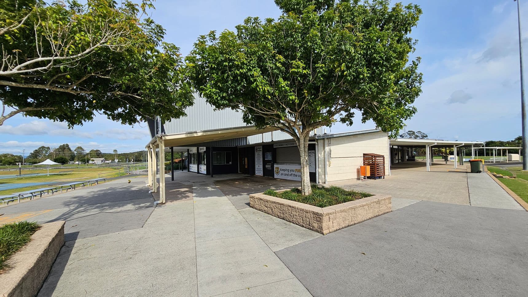 A paved outdoor area with a building in the centre, featuring a covered walkway. Two trees with lush green foliage are planted in raised brick planters. On the left, there are benches and a sports field with a white canopy in the distance. On the right, a pathway leads to more covered areas and a bin is visible. The ground is a mix of grey and green paving.