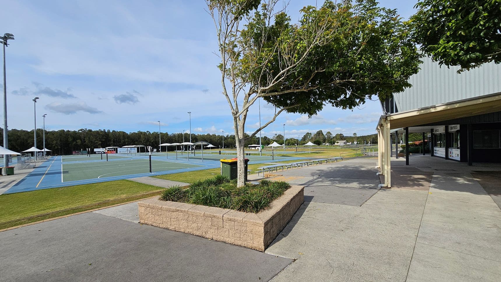 On the left, there are several outdoor sports courts with blue and green surfaces, surrounded by tall floodlights and white tents. In the middle, a tree is planted in a raised brick planter with grass. A green and yellow bin is nearby. On the right, a building with a green roof and a covered walkway is visible. The ground is a mix of concrete and grass, and there are benches along the path. In the background, there are trees and open fields.