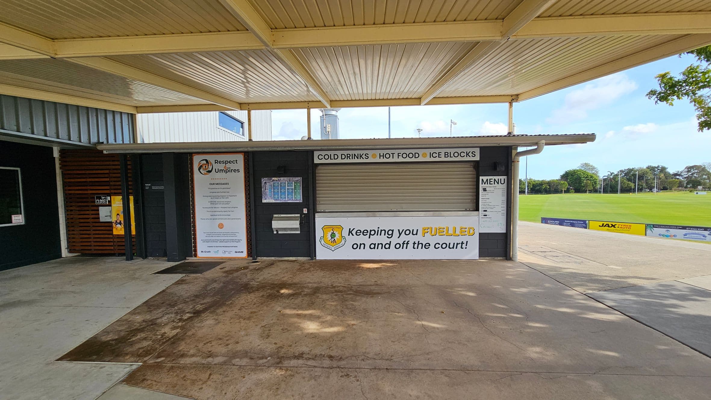A closed kiosk with a corrugated metal roof and a concrete floor. The kiosk has a shuttered window with signs above it reading "COLD DRINKS," "HOT FOOD," and "ICE BLOCKS." A large sign on the left promotes respect for umpires. Another sign below the window says, "Keeping you FUELLED on and off the court!" A menu is displayed on the right. In the background, there is a grassy sports field with trees and a few advertisements along the fence.
