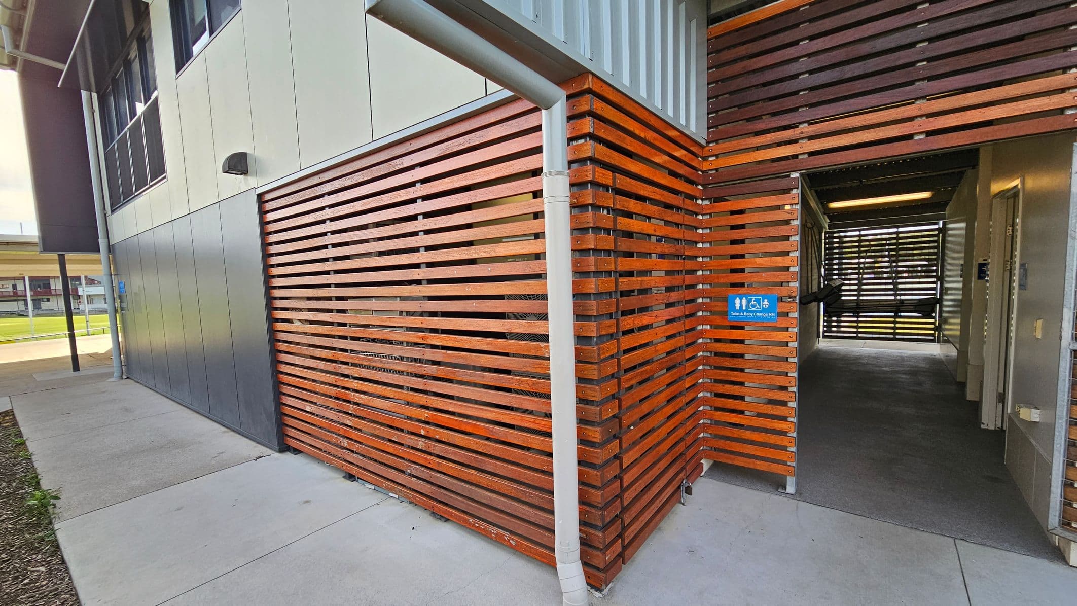 A modern building exterior with a combination of grey metal panels and horizontal wooden slats. The wooden slats form a corner and continue into a passageway. A white downpipe is attached to the corner. The entrance to the passageway has a blue sign indicating accessible toilets and baby changing facilities. The floor is concrete, and the interior passageway has a dark carpet. In the background, there is a glimpse of a grassy area and another building.