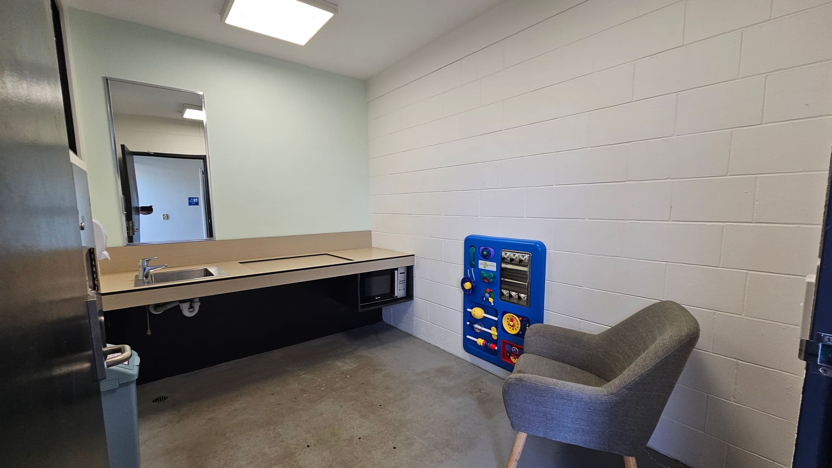 A small room with white brick walls and a grey concrete floor. On the left, there's a countertop with a sink and a microwave underneath. Above the counter is a mirror. On the right, a grey upholstered chair sits next to a blue activity panel attached to the wall. The ceiling has a square fluorescent light fixture.