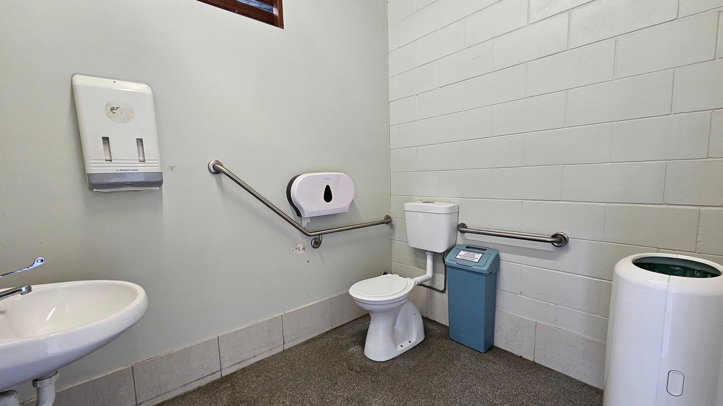 A public toilet with a white toilet bowl and cistern in the centre, equipped with metal grab bars on both sides. To the left, a white sink with a metal tap is visible, along with a wall-mounted paper towel dispenser. On the right, a blue sanitary bin and a white waste bin are present. The walls are tiled in white, and the floor is covered with a mix of grey and brown tiles.