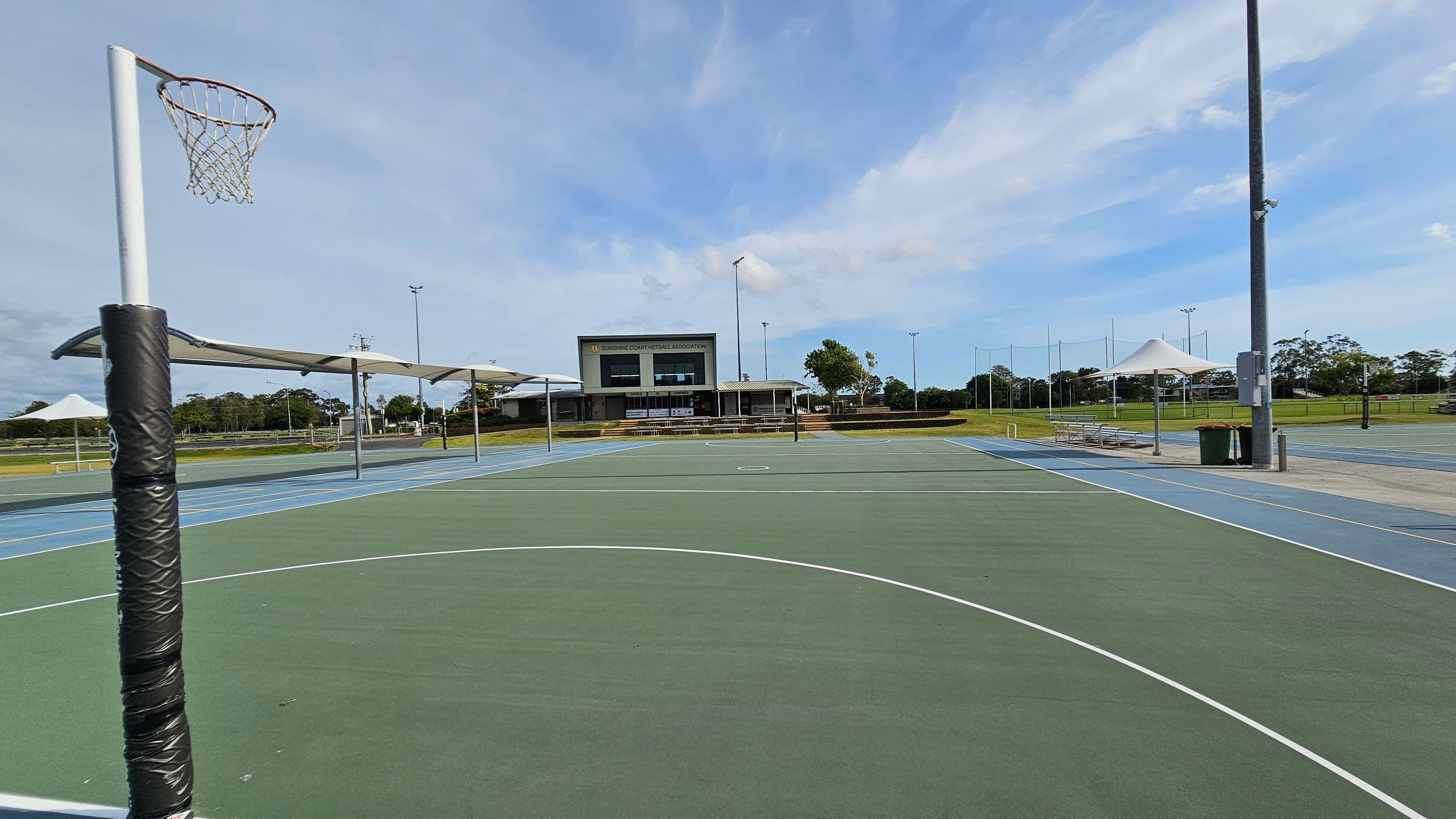Outdoor netball courts with a green playing surface and white lines. A netball hoop with a padded post is on the left. In the background, there is a building with a sign, "Sunshine Coast Netball Association." Several covered seating areas and a rubbish bin are visible on the right. The area is surrounded by grass and trees, with tall light poles around the courts.