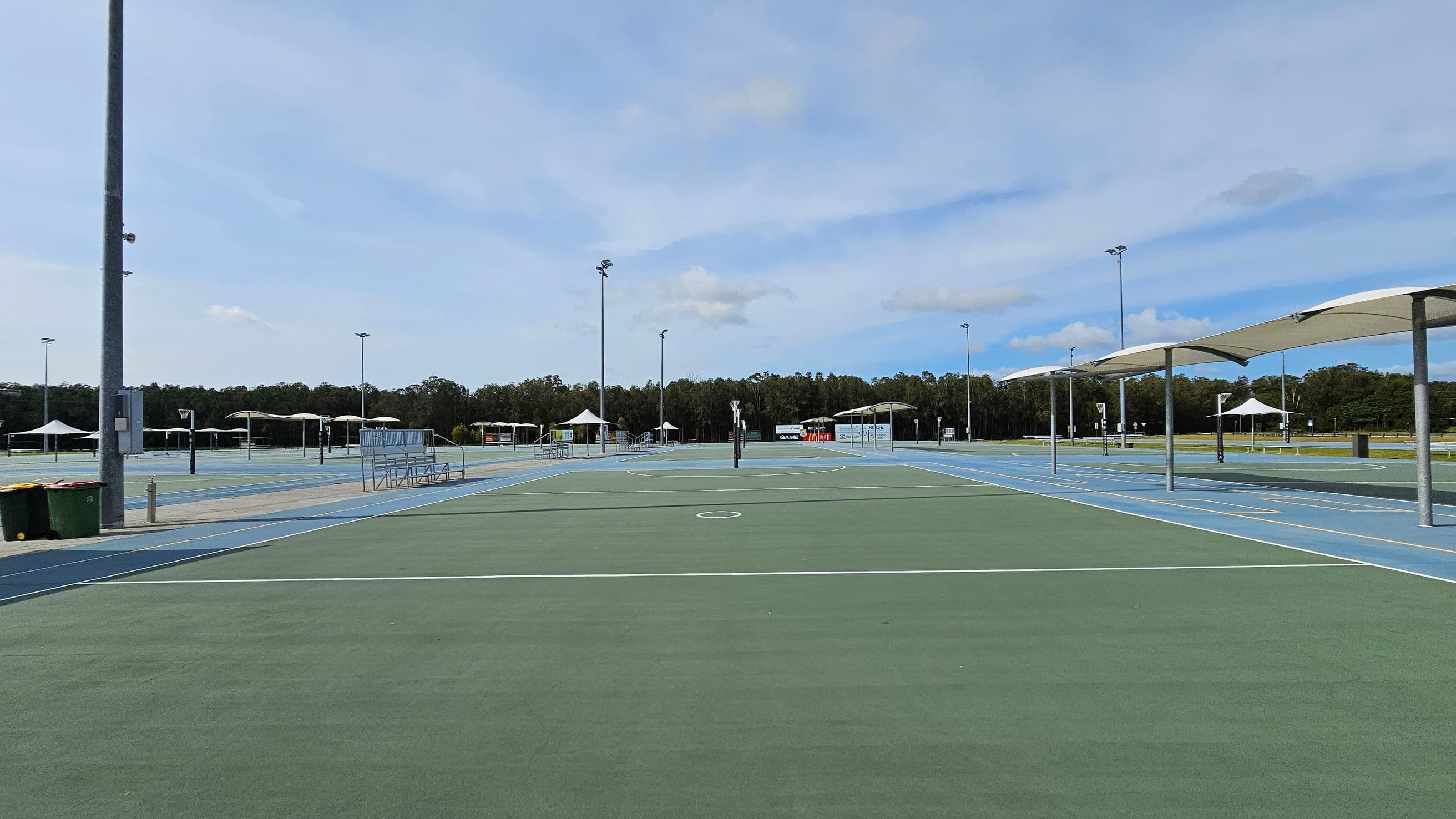 A large outdoor sports complex with multiple netball courts. The courts have green and blue surfaces with white lines. On the left, there are bins and a tall light pole. In the centre, a netball post is visible. To the right, there are covered seating areas with white canopies. In the background, there are more courts, additional canopies, and a line of trees.