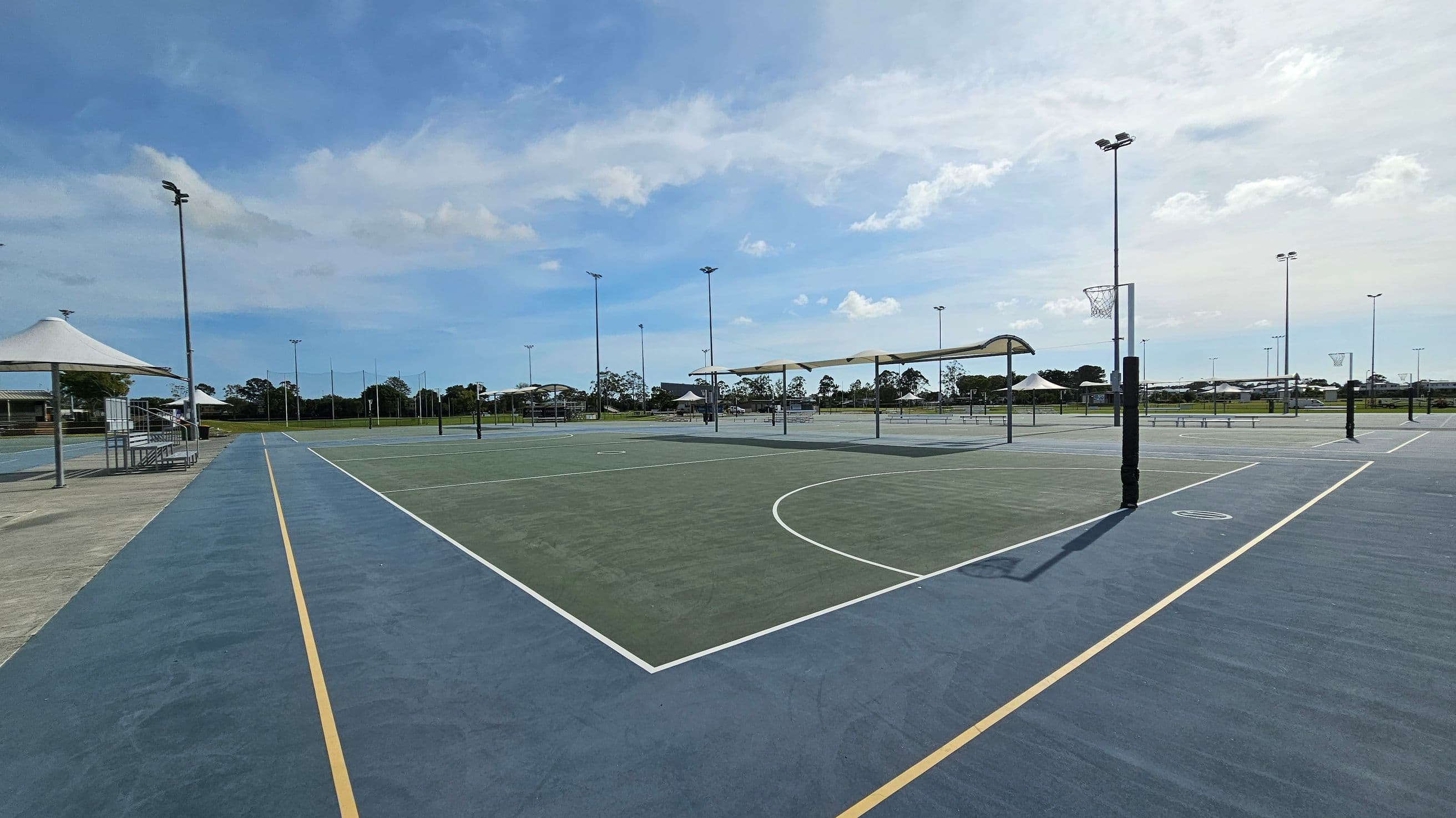 Outdoor netball courts with blue and green surfaces, marked with yellow and white lines. On the left, a covered seating area with a white canopy and metal benches. Several tall floodlights are positioned around the courts. In the background, there are more courts and some trees. The area is open and spacious, with clear visibility of the surrounding landscape.