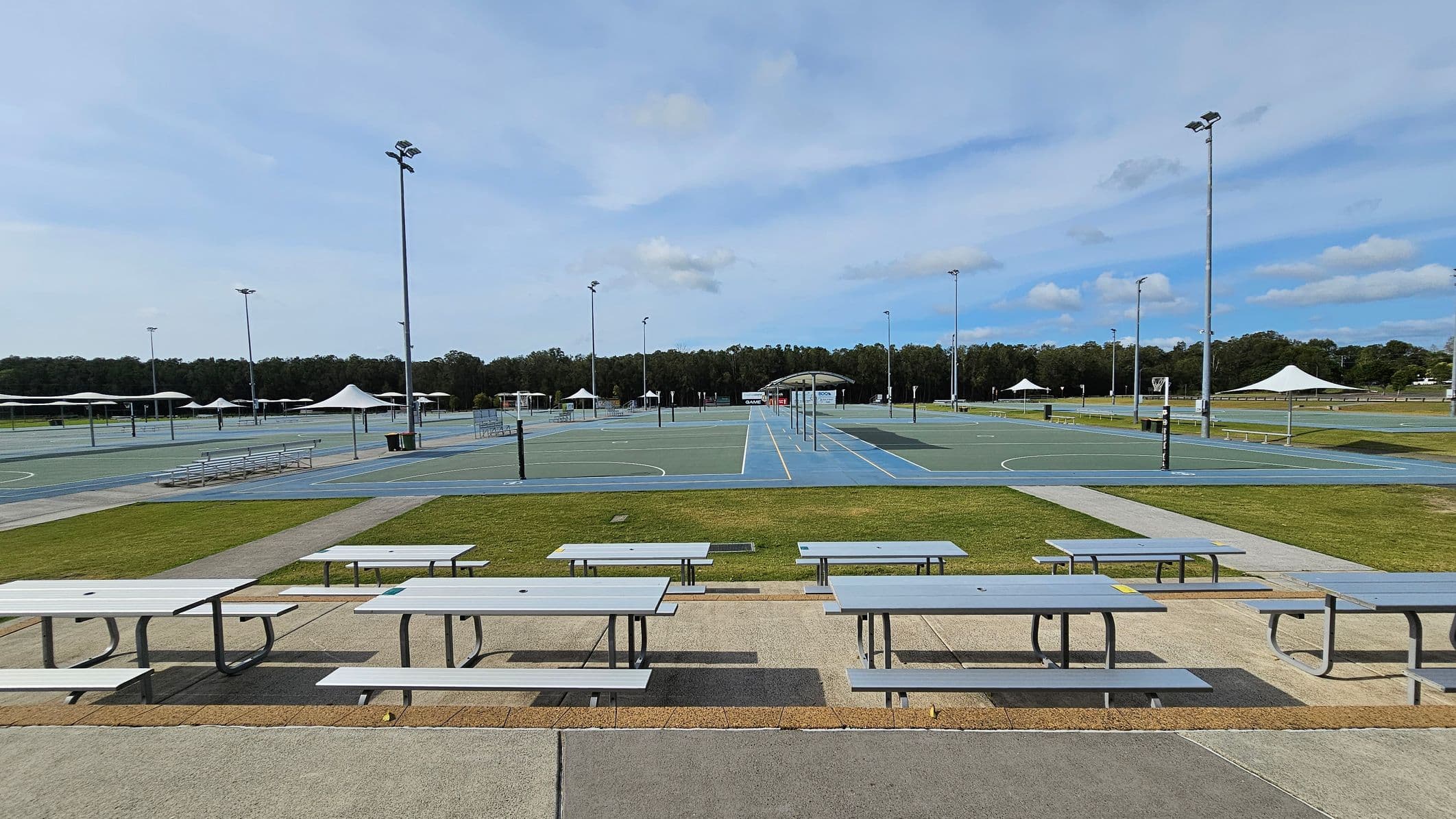 Rows of light grey picnic tables are in the foreground on a paved area. Beyond them, there are multiple outdoor sports courts with blue and green surfaces, surrounded by tall floodlights. White shade structures are scattered around the courts. In the background, there is a line of trees bordering the area. The ground is a mix of grass and concrete paths.