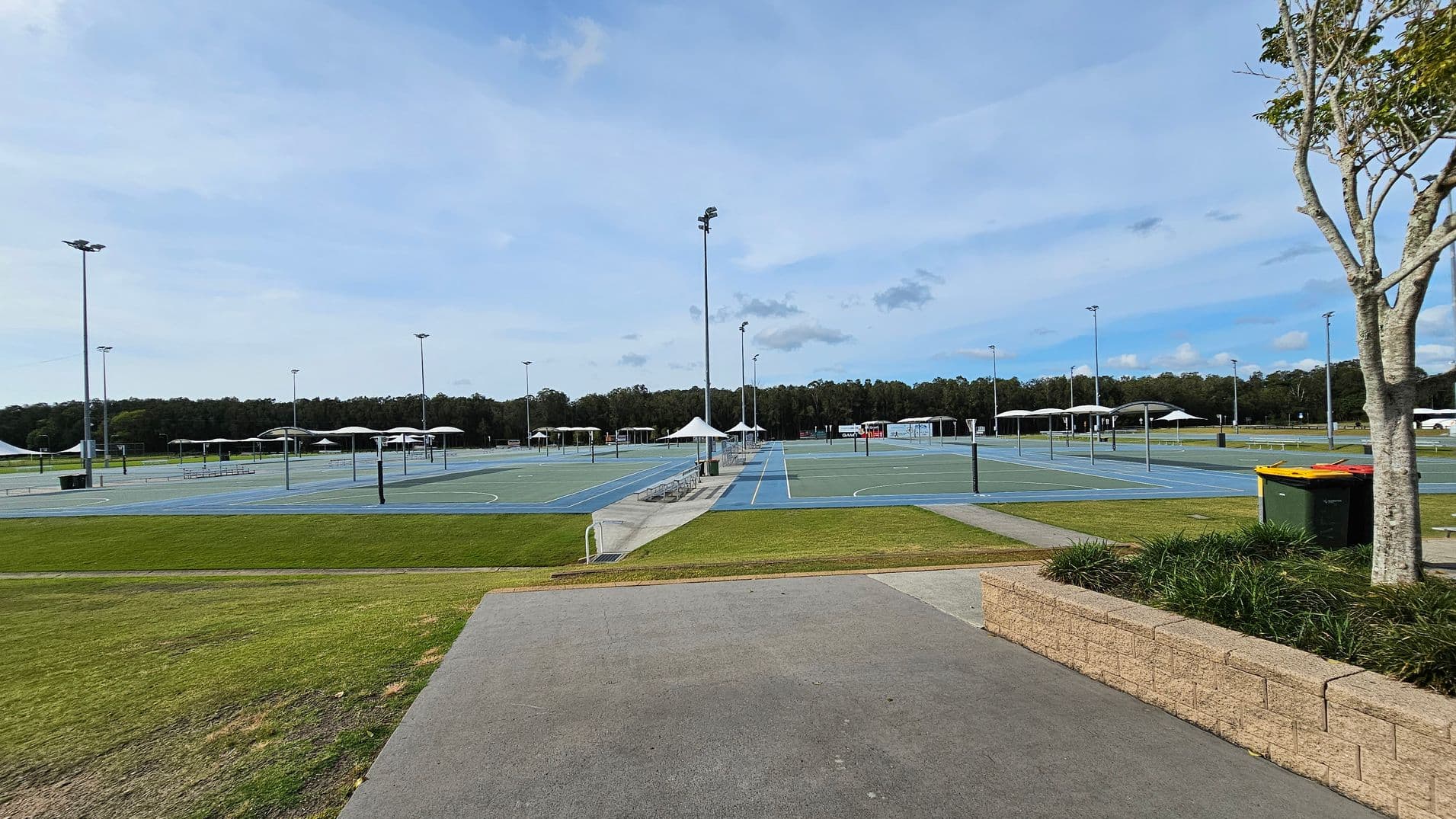 A series of outdoor sports courts with blue surfaces and white lines, surrounded by tall floodlights. In the background, there are several white canopies providing shade. The foreground features a paved path and a grassy area with a low brick wall on the right. A tree and a green rubbish bin with a yellow lid are also on the right. Dense trees form a line in the distant background.