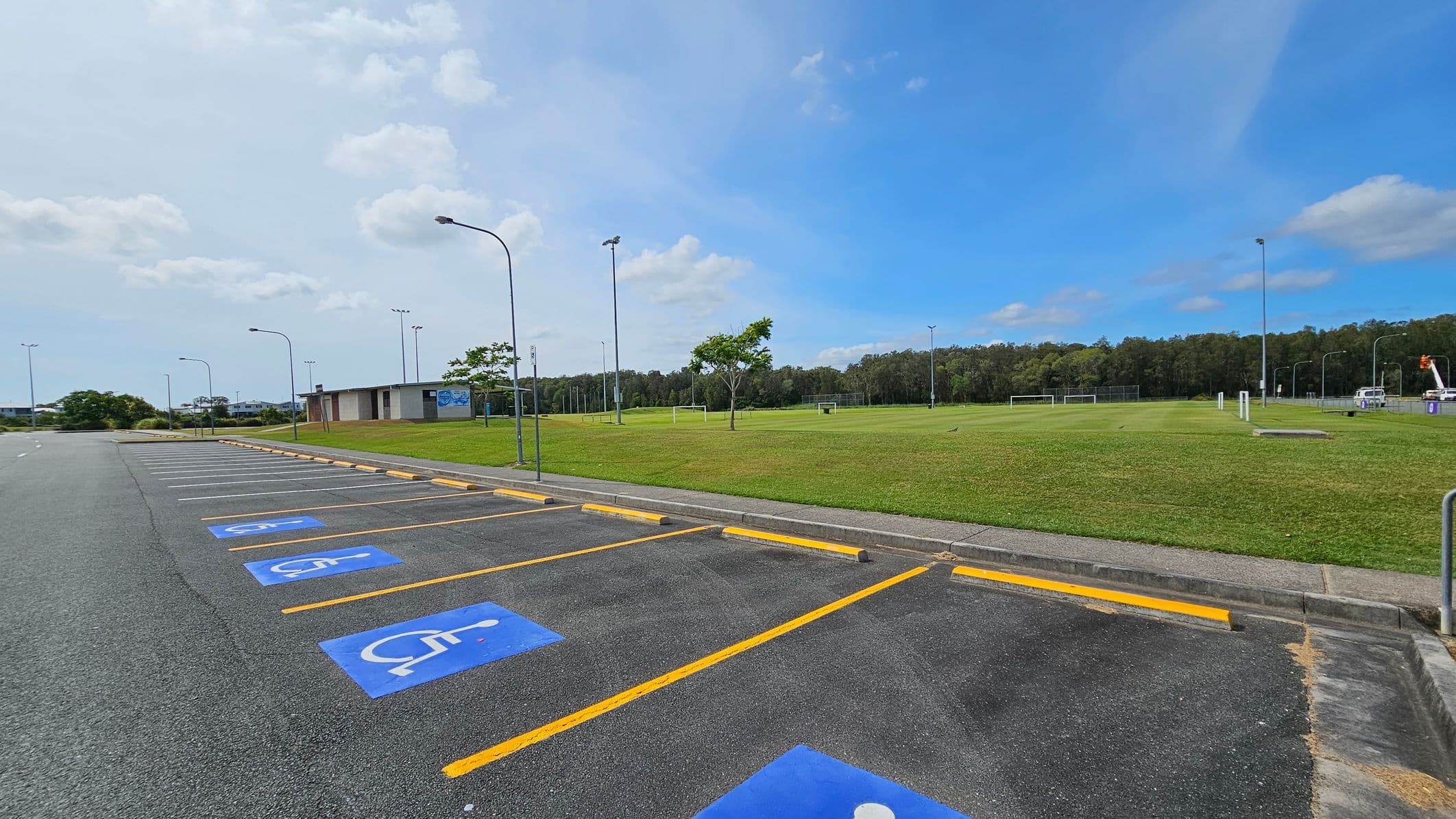 A parking area with several marked disabled parking spaces on the left, featuring blue symbols on the asphalt. Yellow lines separate the spaces. On the right, there is a large grassy field with a few trees and football goals. A small building is visible in the background, along with tall light poles. The area is bordered by a line of trees in the distance.