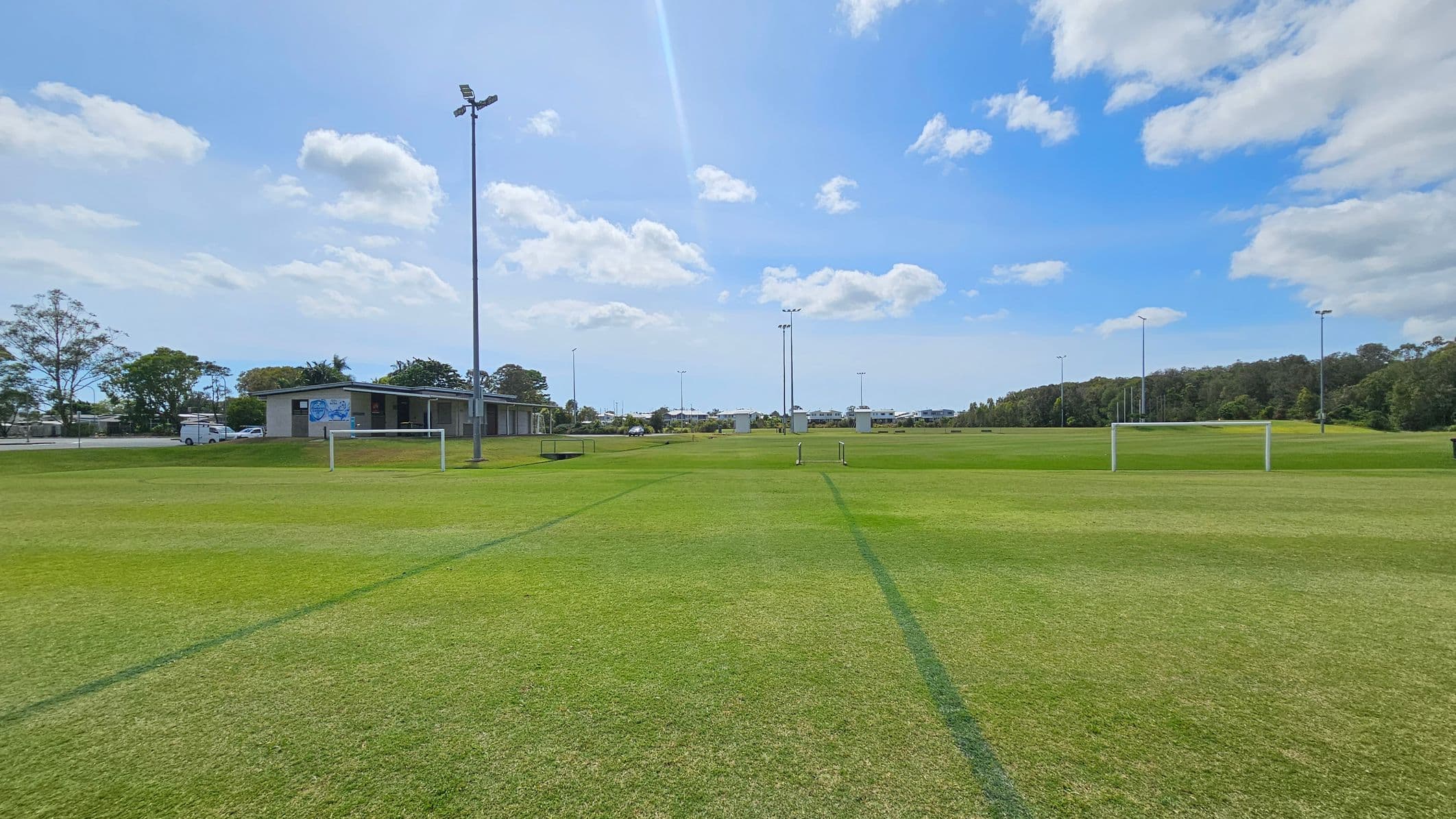 A grassy sports field with goalposts in the foreground. In the background, there is a small building to the left, with a few parked vehicles nearby. Several tall floodlights are scattered across the field. Trees line the horizon on the right side, and a few structures are visible in the distance. The sky is partly cloudy.