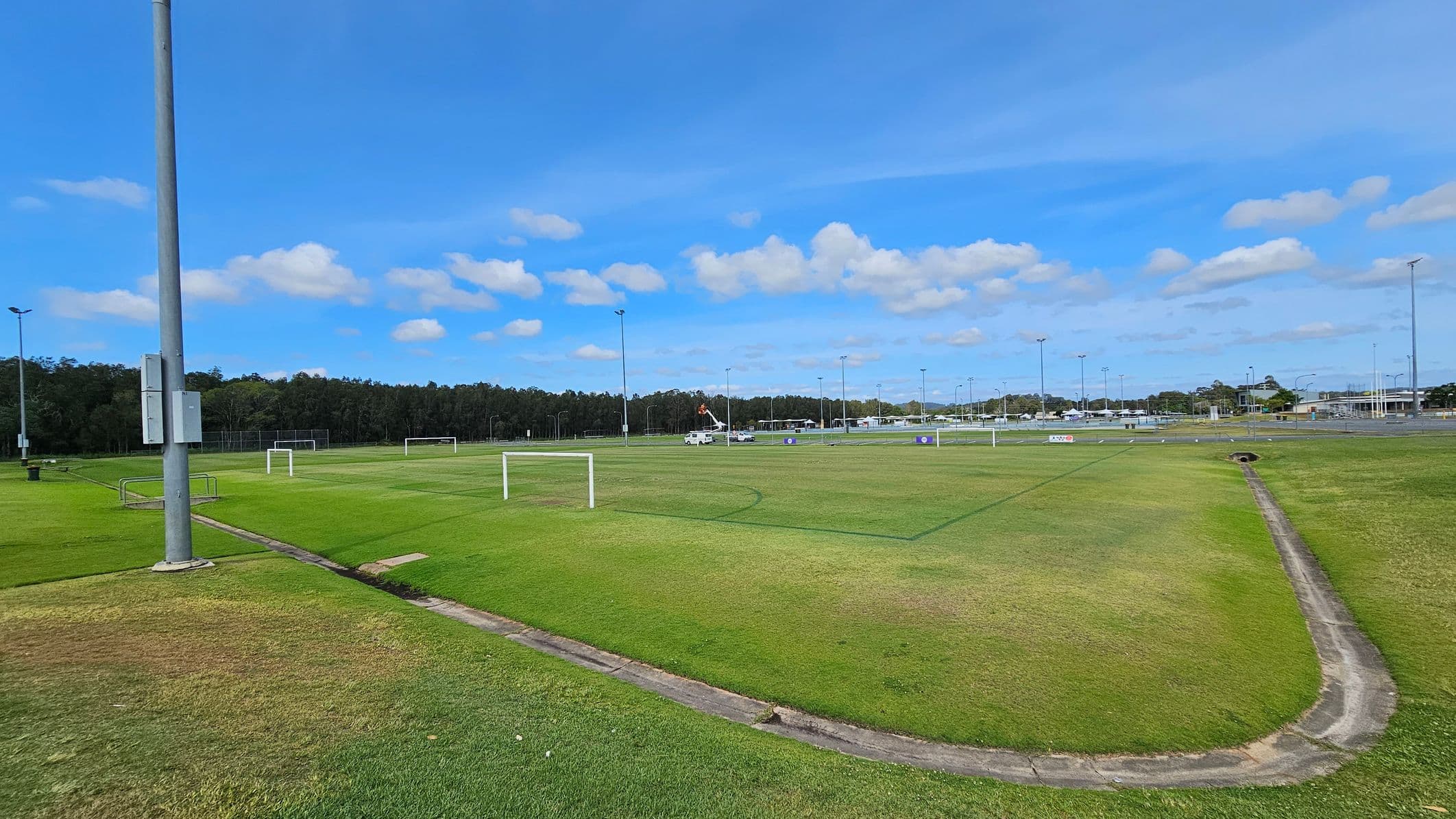 A large grassy sports field with several white goalposts scattered across it. On the left, a tall floodlight pole stands near the edge of the field. In the background, a line of trees borders the field, and a vehicle with a raised platform is visible. To the right, there are more floodlights and a few buildings in the distance. The ground is a mix of green and brown patches, with a narrow concrete drainage channel running through it.