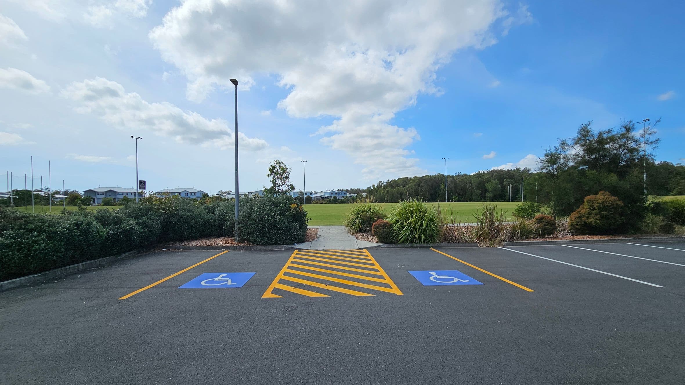 Two parking spaces with blue disabled symbols are marked on the asphalt, separated by yellow diagonal lines. A path leads from the parking area through low bushes and greenery towards a grassy field in the background. A tall lamp post stands near the path. In the distance, there are houses and trees lining the horizon.