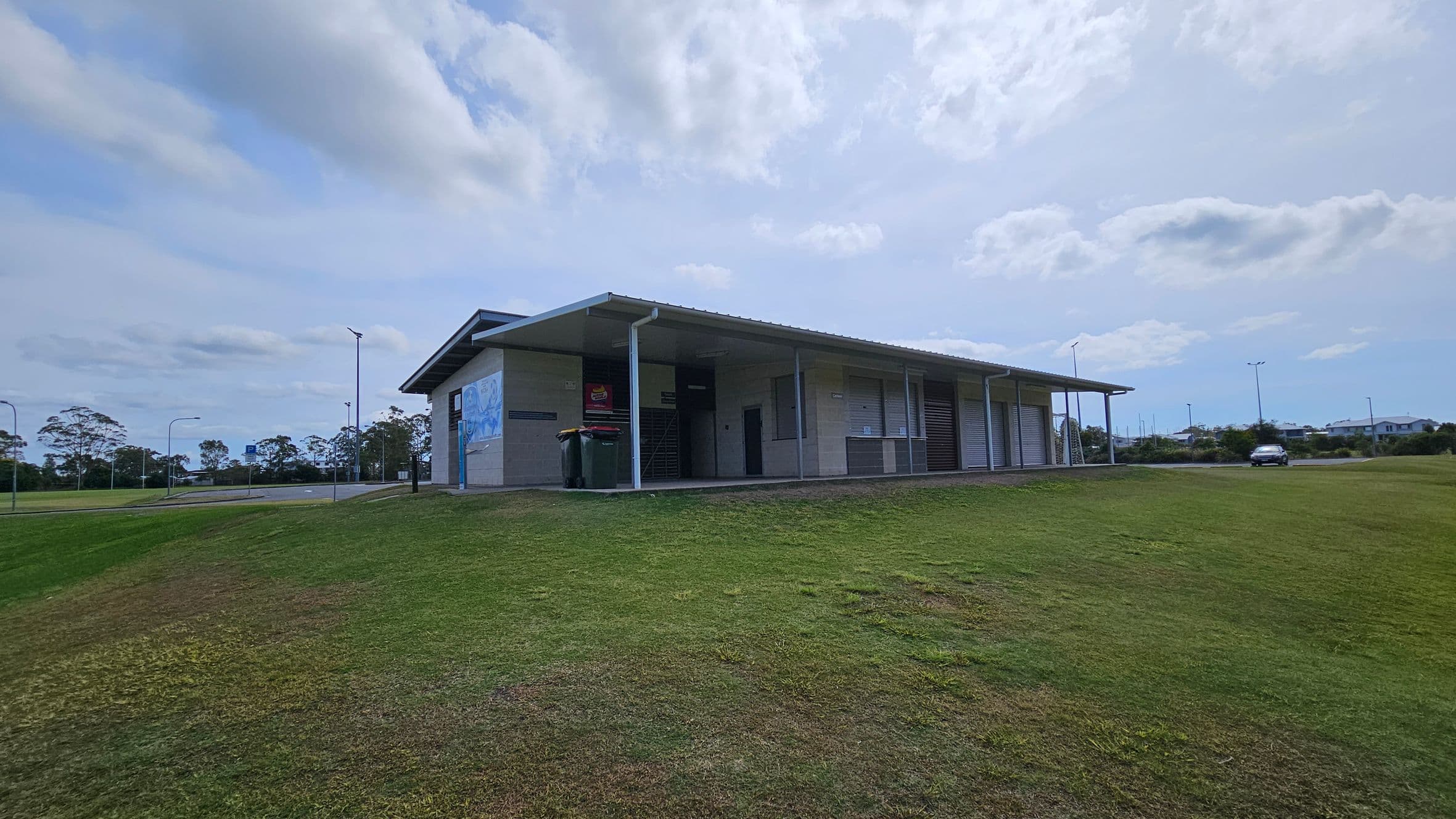 A single-storey building with a sloped roof is situated on a grassy hill. The structure has a covered walkway with several roller doors and a mural on the left wall. Two bins are placed near the entrance. In the background, there are trees and a few streetlights. A car is parked on the right side, near a road that runs alongside the building.