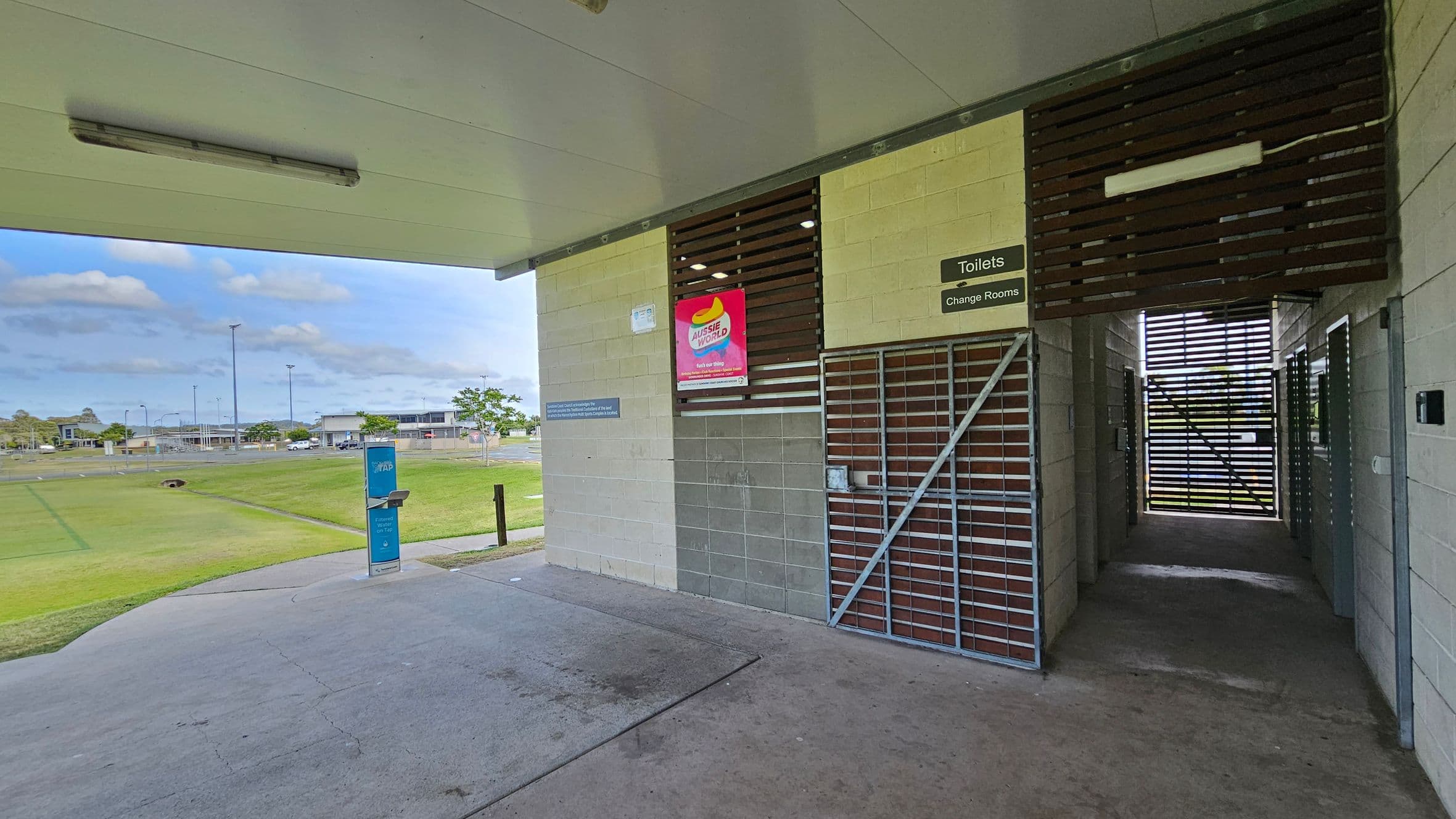 A sports facility with a covered area. On the right, there is a gated entrance to toilets and change rooms, with a sign above. A poster is displayed on the wall. The floor is concrete. On the left, an open grassy field is visible, with a water fountain in the foreground. In the background, there are buildings and a car park. The ceiling is flat with a light fixture.